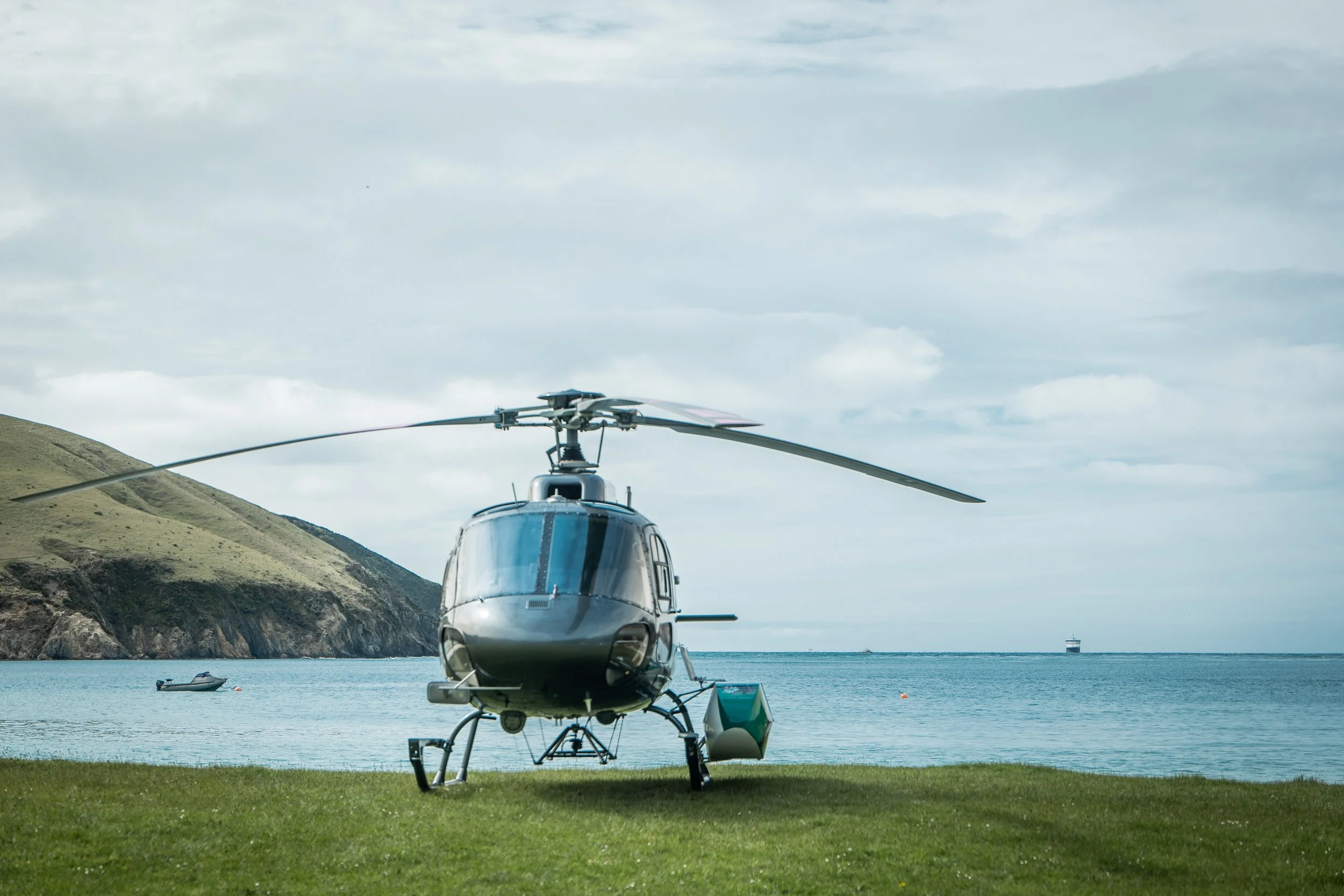 A helicopter parked on a grassy area near the ocean, with hills in the background and a boat floating on the water.