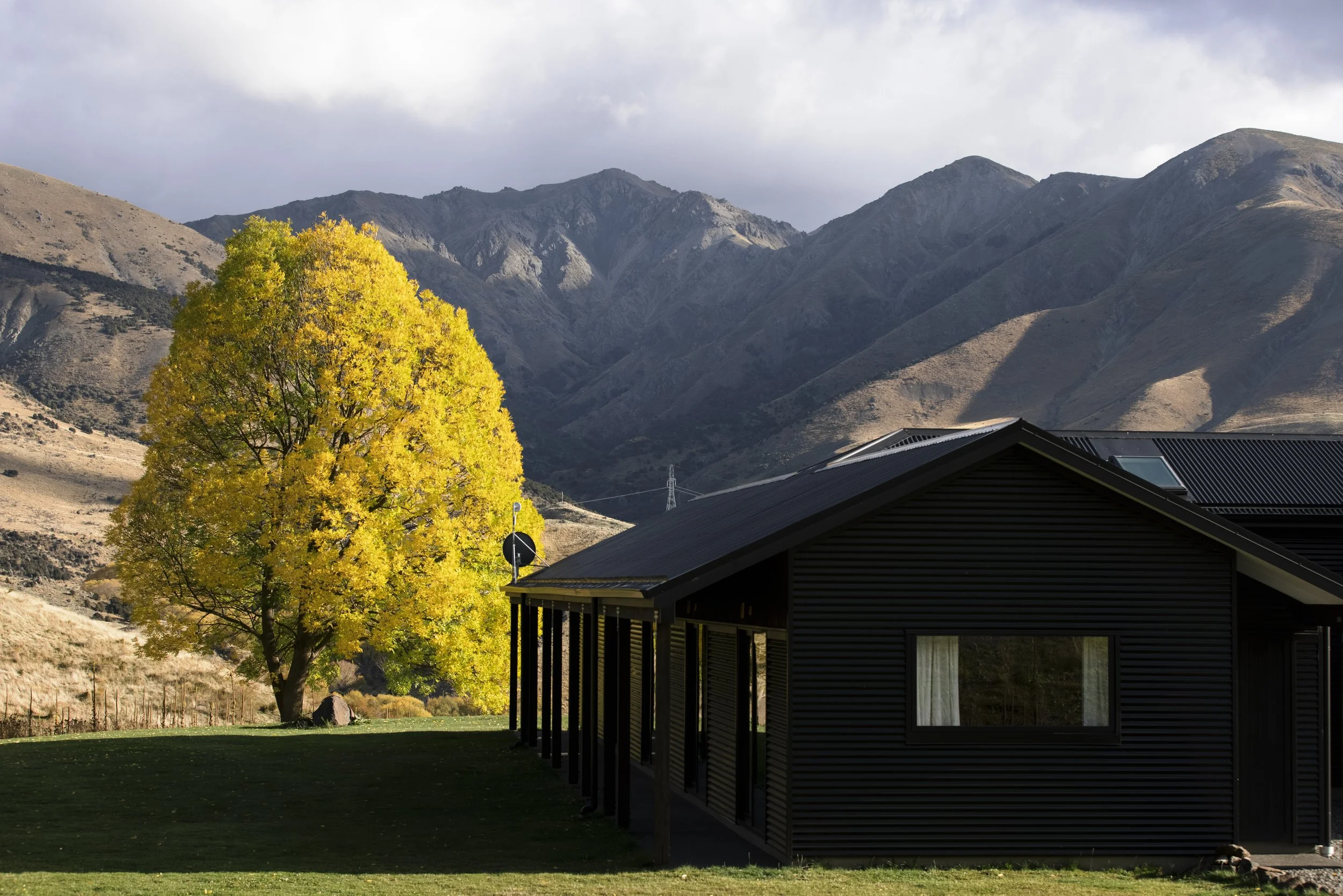 A house with a black roof and covered porch in a grassy yard, a yellow-leaved tree, rugged mountains in the background, and cloudy sky.