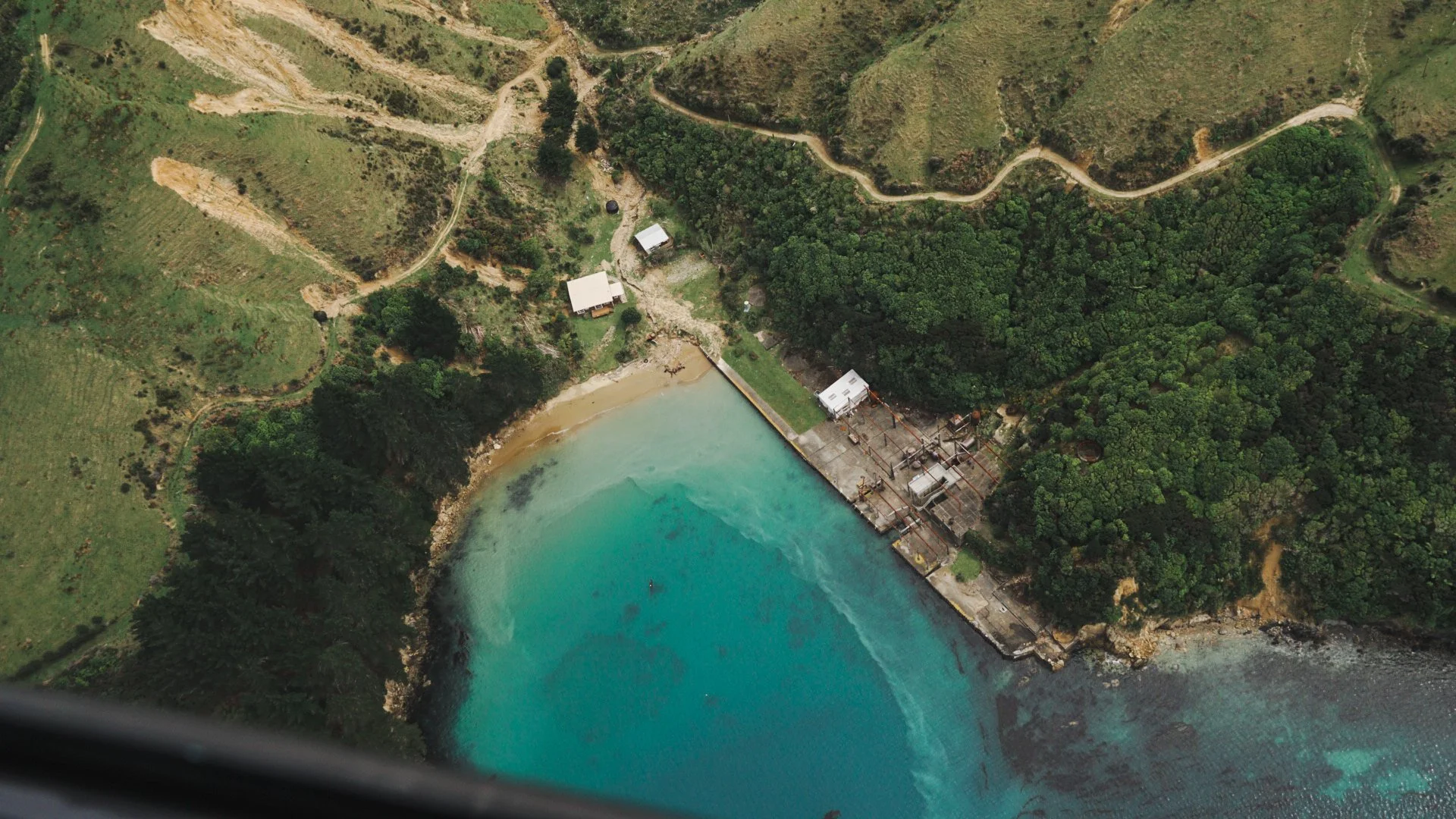 Aerial view of a dam on a coastline with turquoise water, surrounded by green hills and forest, and a few buildings nearby.