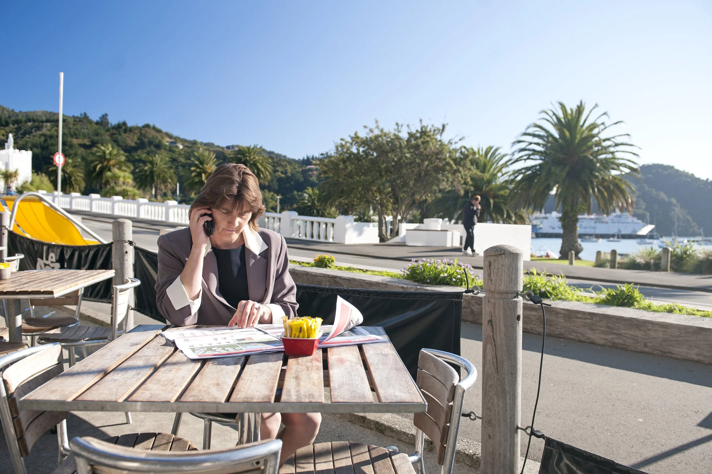 Woman sitting at an outdoor restaurant table talking on a cell phone, with a harbor, palm trees, and a cruise ship in the background on a sunny day.