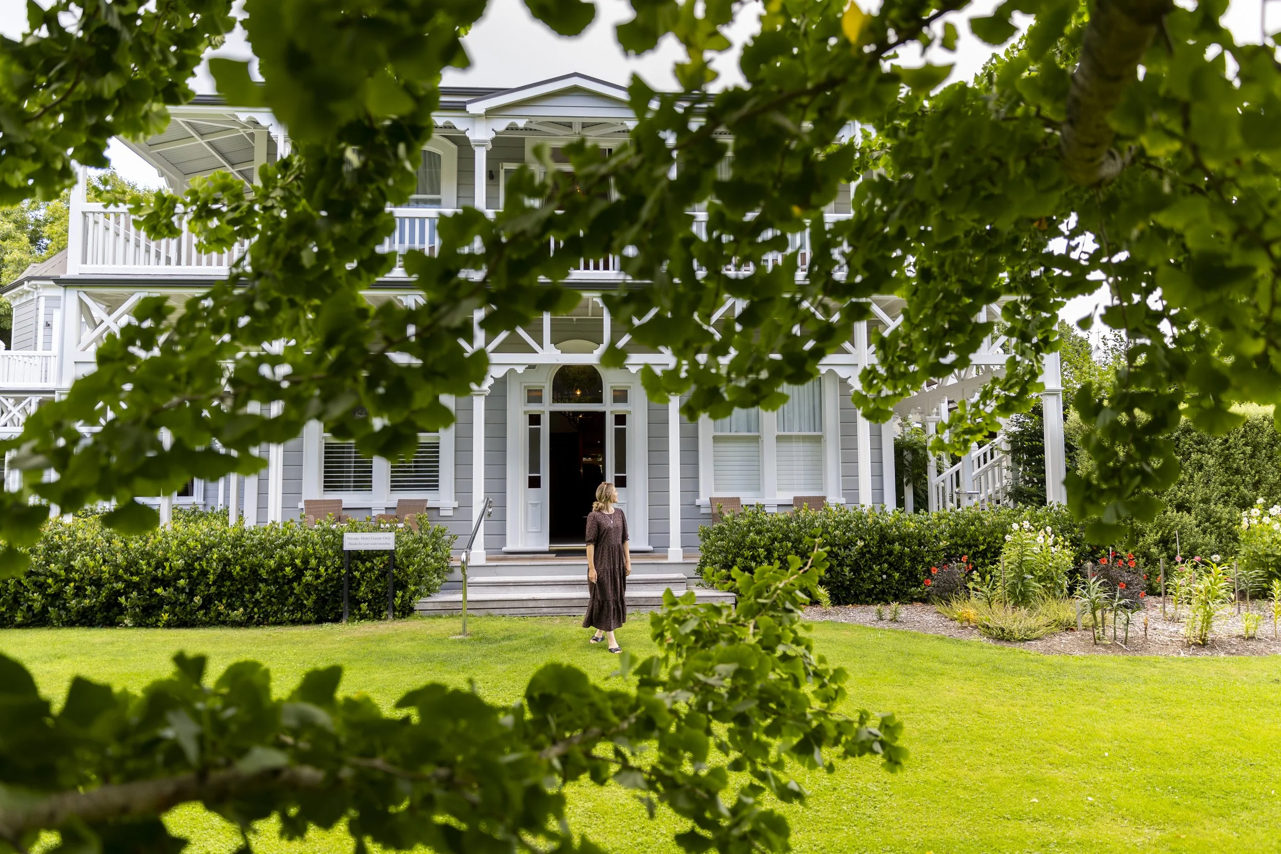 A woman in a brown dress standing on a grassy lawn in front of a white, two-story house with a porch, surrounded by greenery and colorful flowers, viewed through a frame of leaves.
