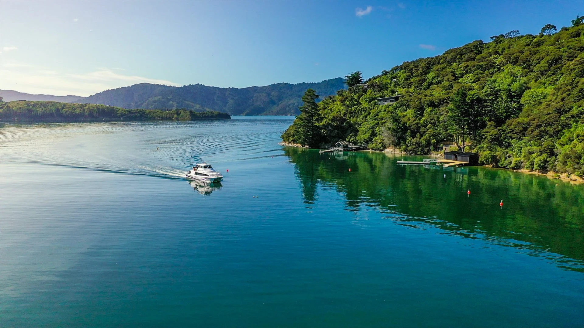 A boat sailing on a calm, greenish-blue lake surrounded by lush, green hills under a clear blue sky.