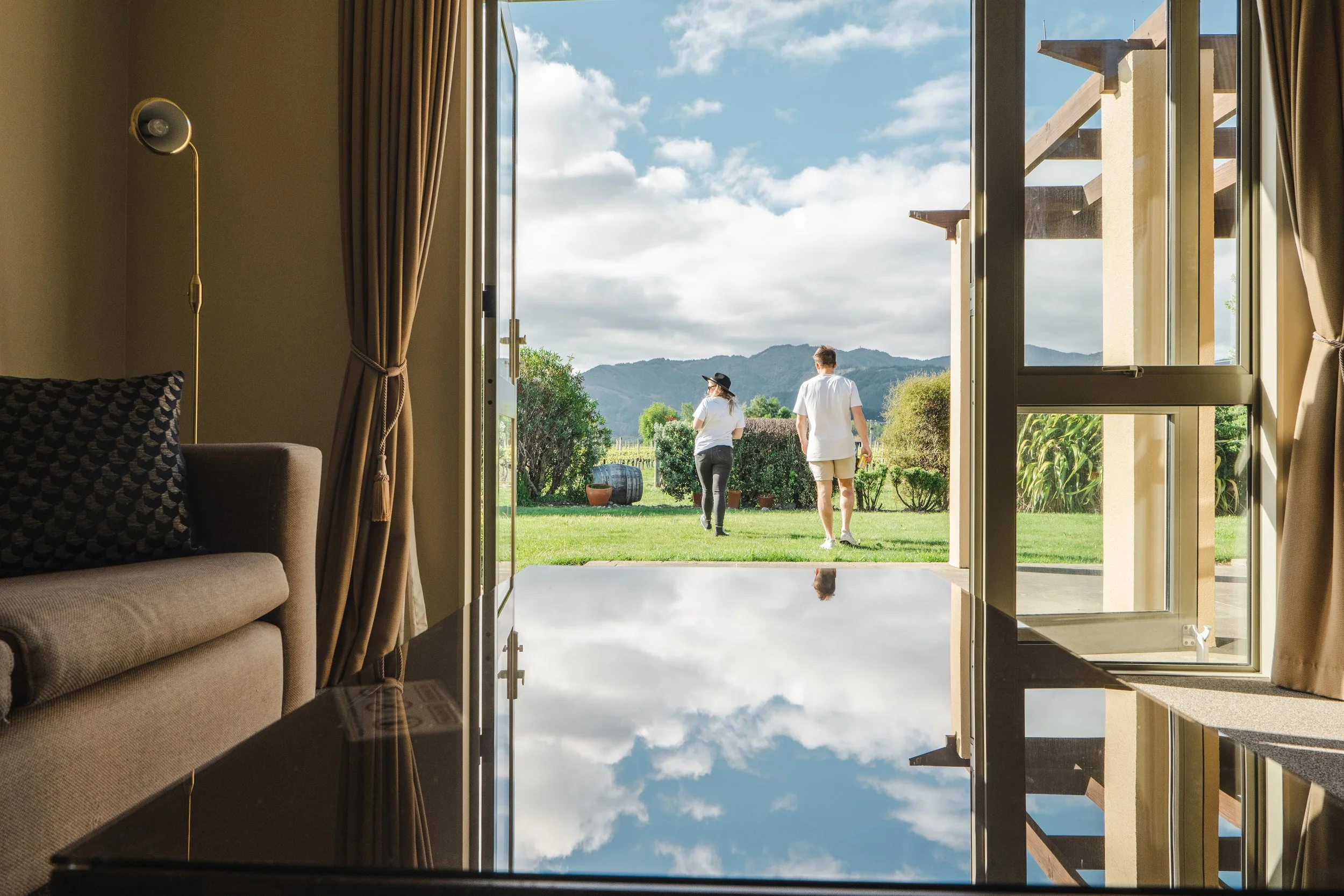 View from inside a room looking out through sliding glass doors onto a garden with two people walking away toward distant mountains.