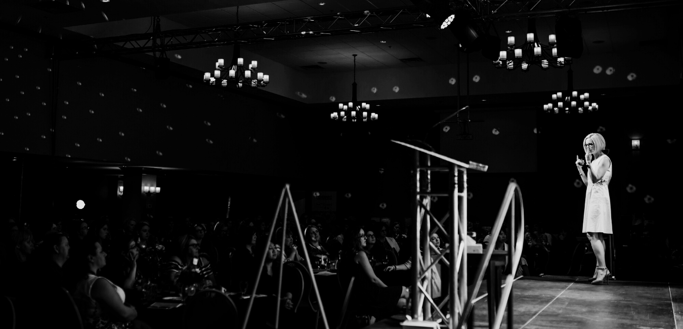 Claire Von Cleveland wearing glasses and a white dress, standing on a stage, speaking into a microphone at a conference or event, with an audience seated in front of her.