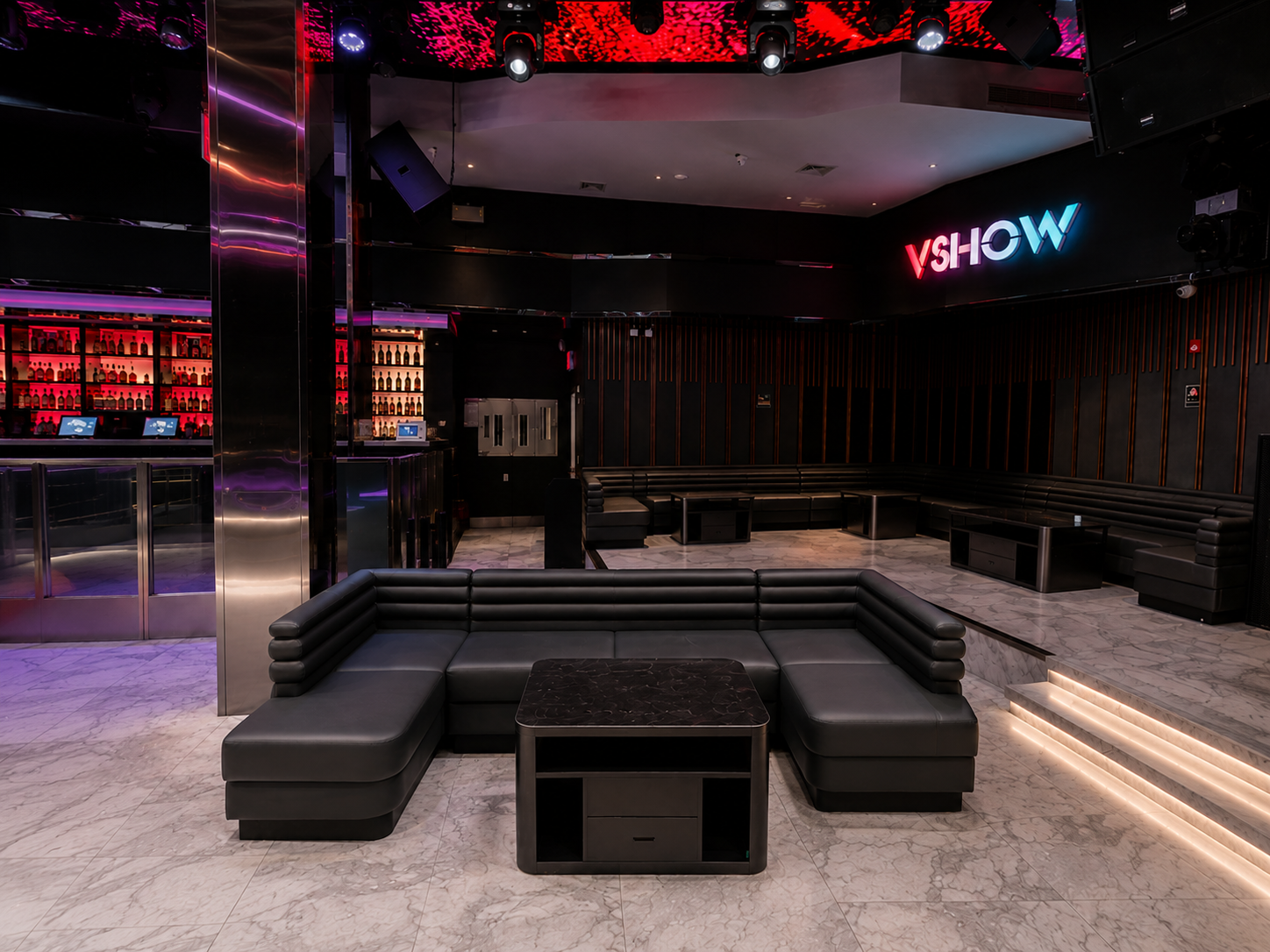 Empty modern lounge with black leather sectional sofas, small tables, a bar with illuminated shelves of bottles, and a sign that says "VSHOW" in colorful neon lights.