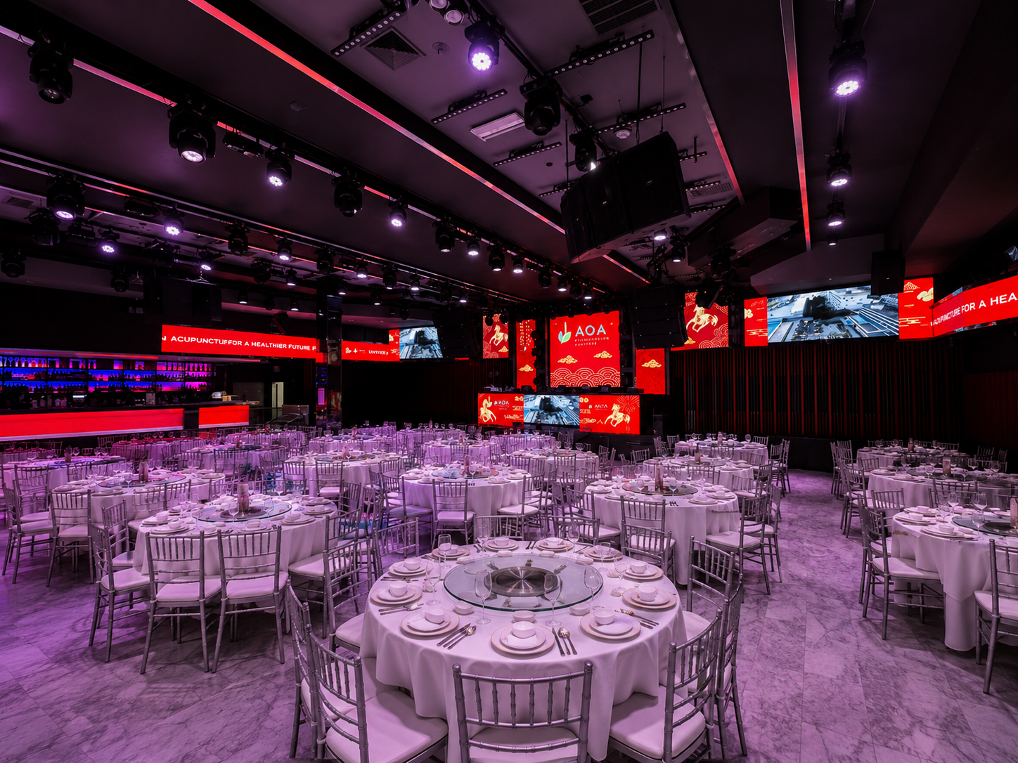 Empty banquet hall with round tables covered in white tablecloths, set for a formal event, in front of a stage with large screens and red lighting.