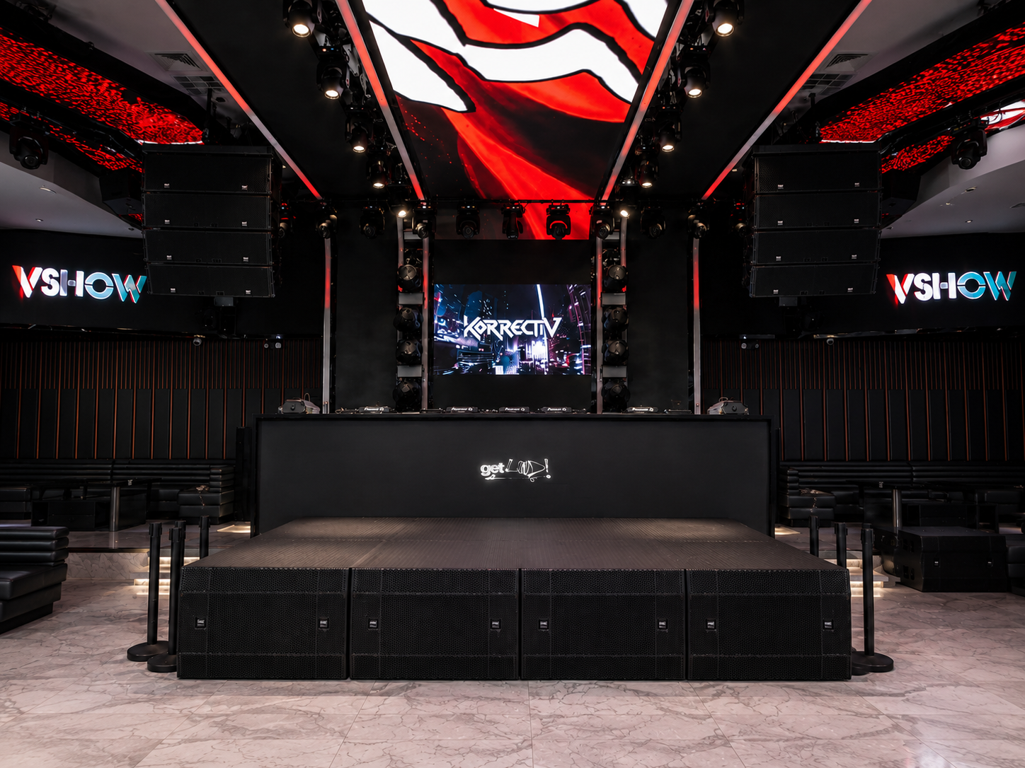 Empty nightclub stage with DJ equipment, large speakers, and digital screens displaying graphics and text including 'KORRECTIV' and 'VSHOW'.