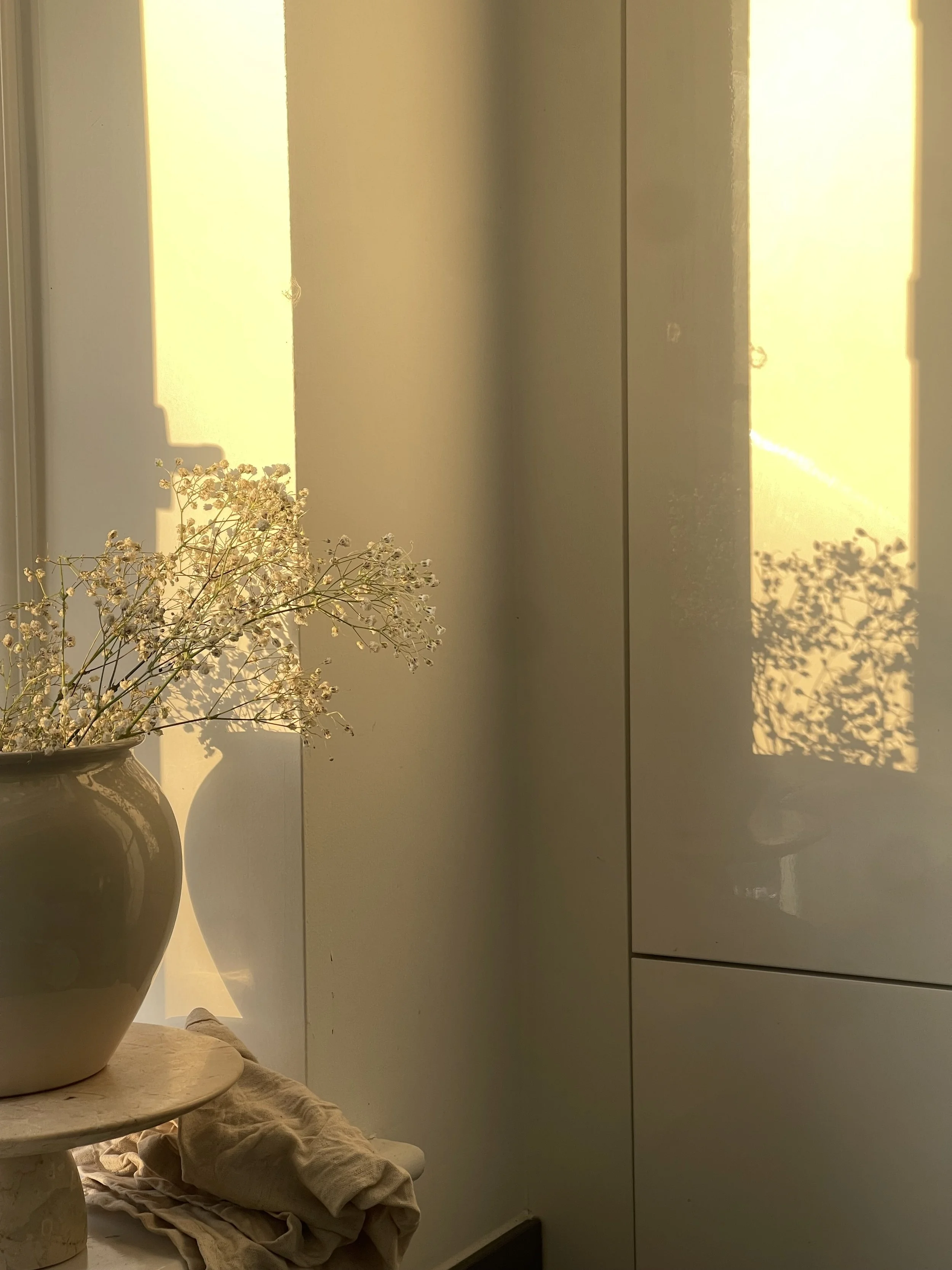 A beige vase with a round opening and a narrow neck holding dried white baby's breath flowers on a small round table. Sunlight casts geometric shadows on a white wall behind them.