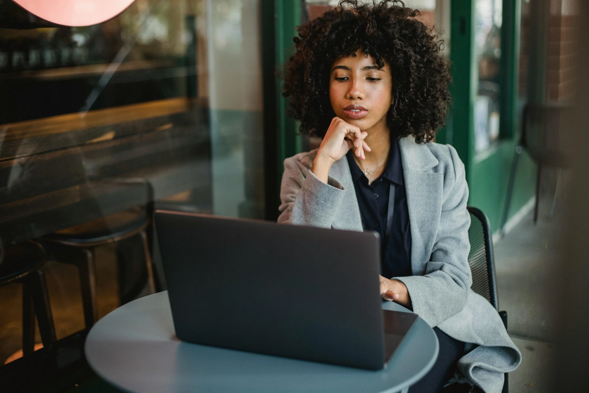 A woman with curly hair wearing a gray blazer and black shirt, sitting at a table in a cafe or restaurant, looking at a laptop screen with a thoughtful expression.
