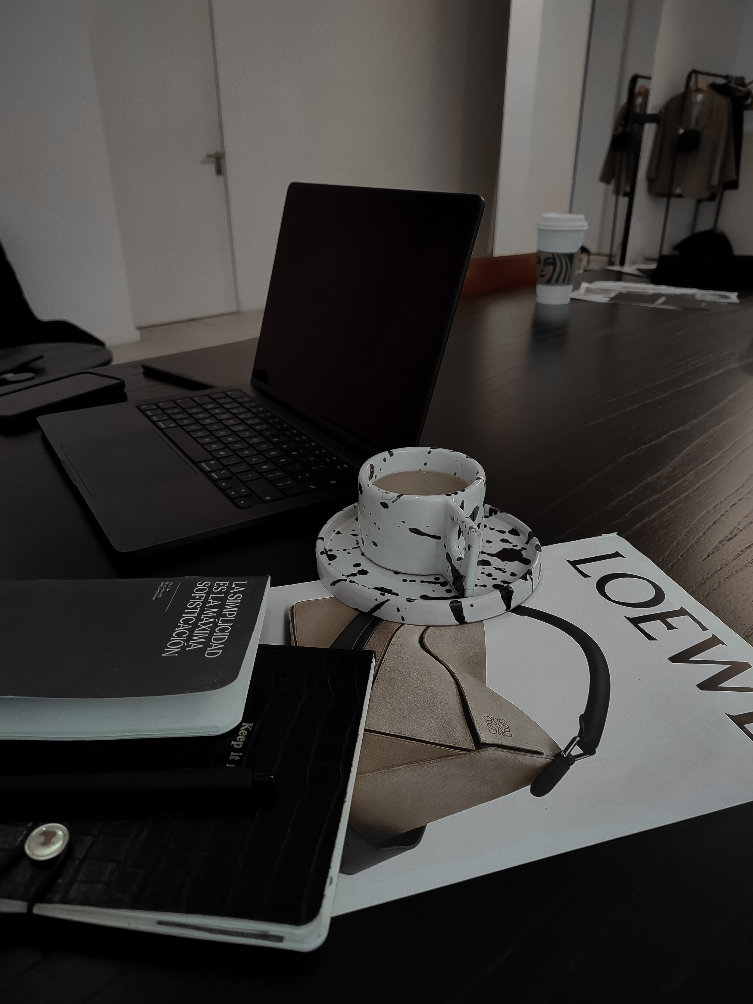 A dark wooden desk with a laptop, a Marie Kondo book, a white ceramic coffee cup with black paint splatters on a matching saucer, a beige purse, and a magazine titled "LOEWE". In the background, there is a closed white door and a coffee cup, papers, and clothing on a rack.
