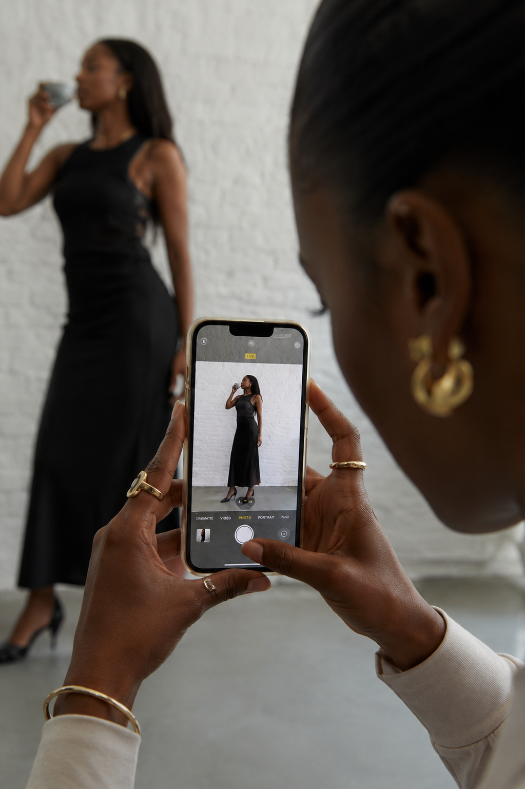 A person taking a photo of a woman in a black dress with a smartphone against a white brick wall.