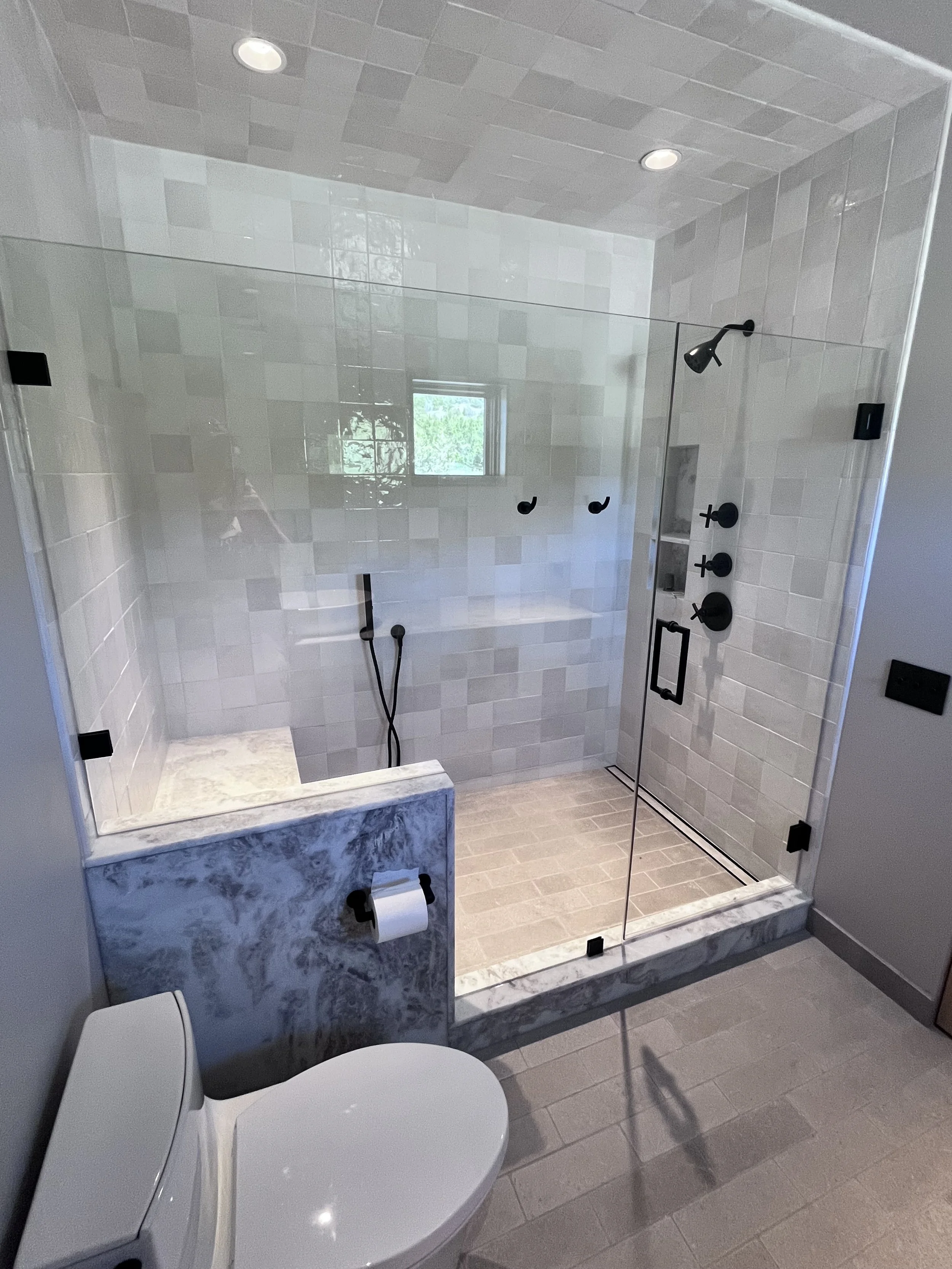 A modern bathroom with a walk-in shower enclosed by clear glass, featuring black fixtures, a marble bench, and beige tiled walls and floor, with a window providing natural light.