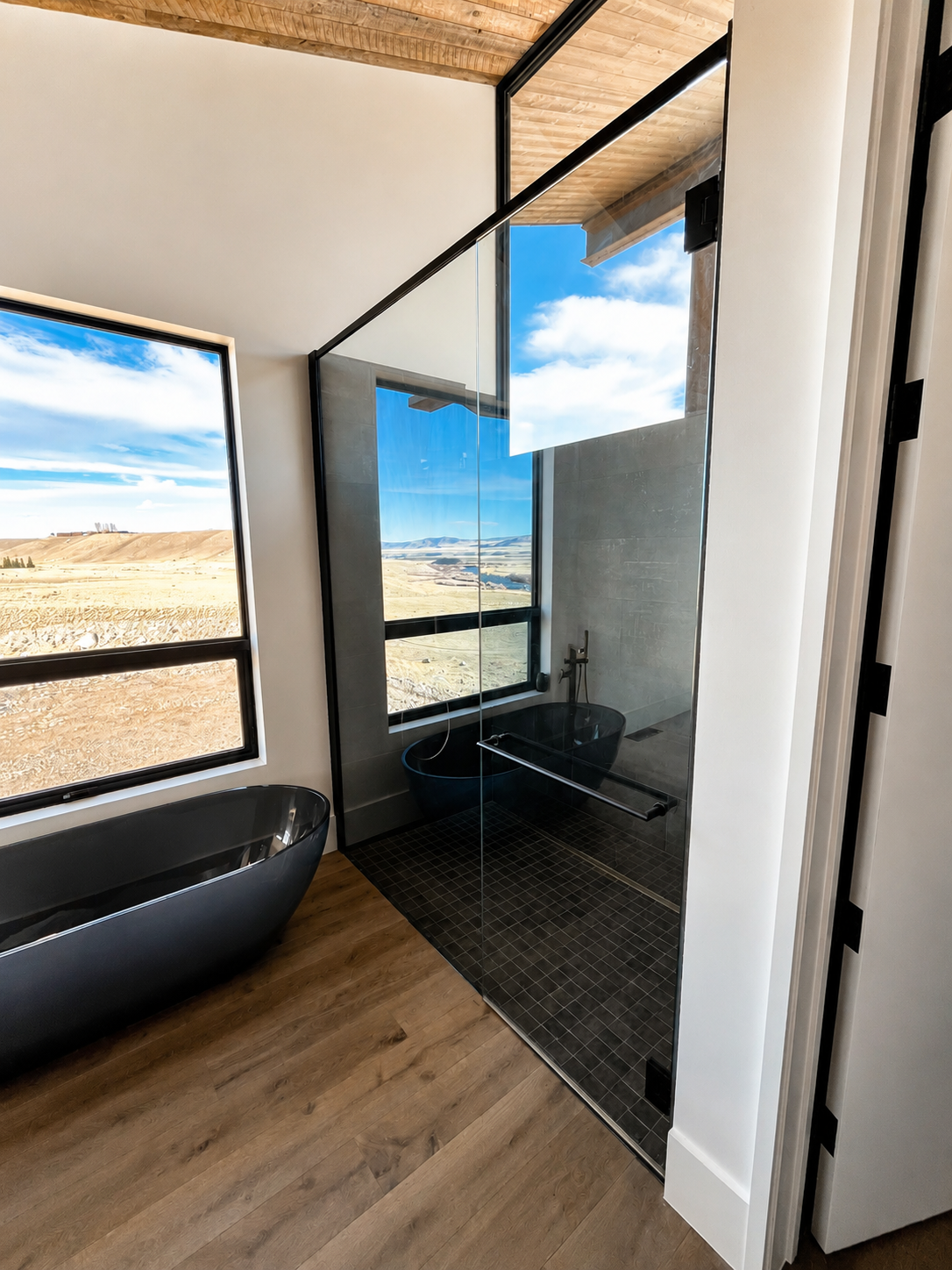 Modern bathroom featuring a black free-standing tub, a walk-in shower with black fixtures and glass walls, and large windows showing a scenic landscape with rolling hills and a partly cloudy sky.