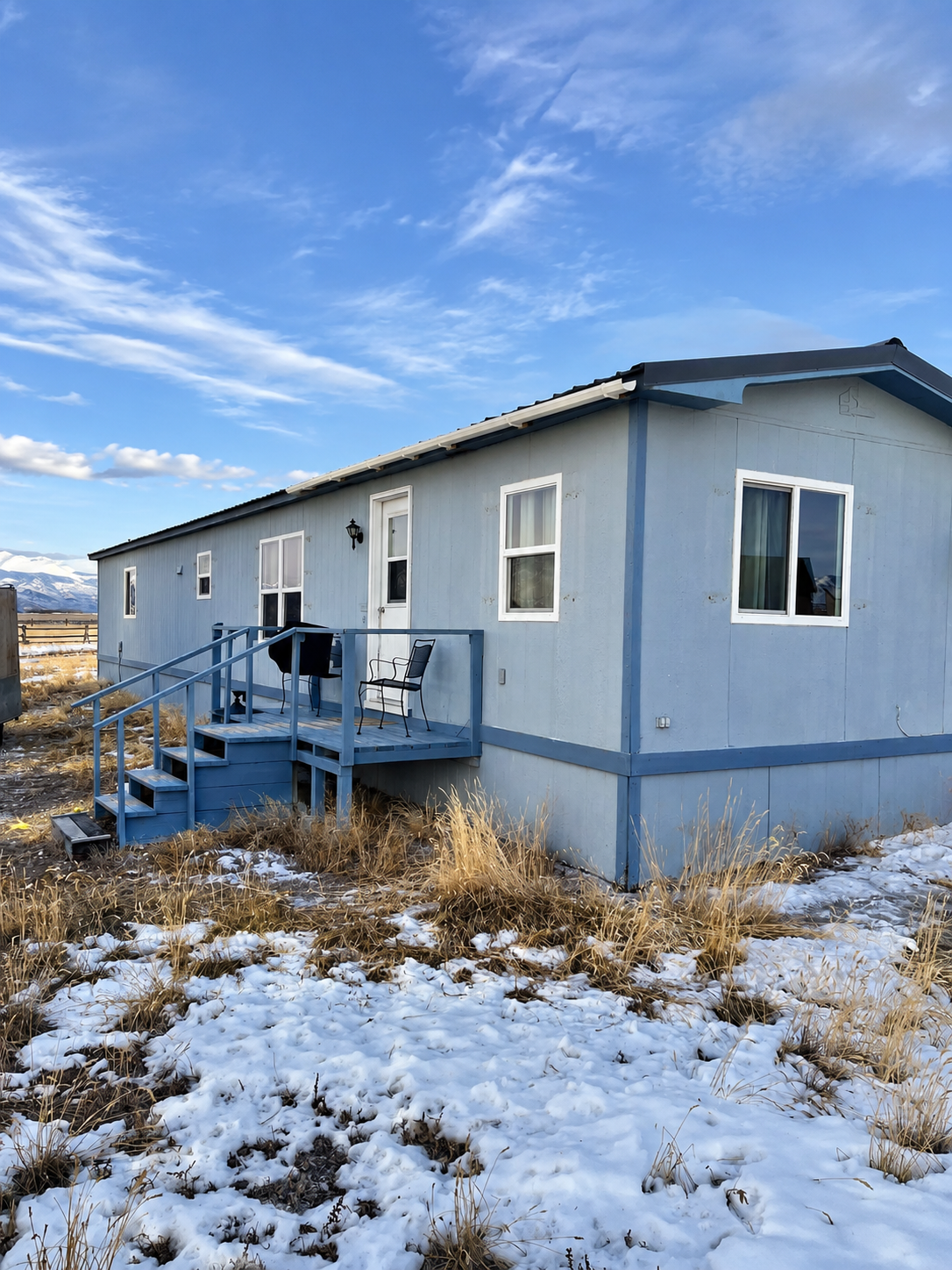 A light blue manufactured home with a small wooden porch, black mailbox, and two chairs outdoors on a partially snow-covered grassy field under a blue sky with scattered clouds.