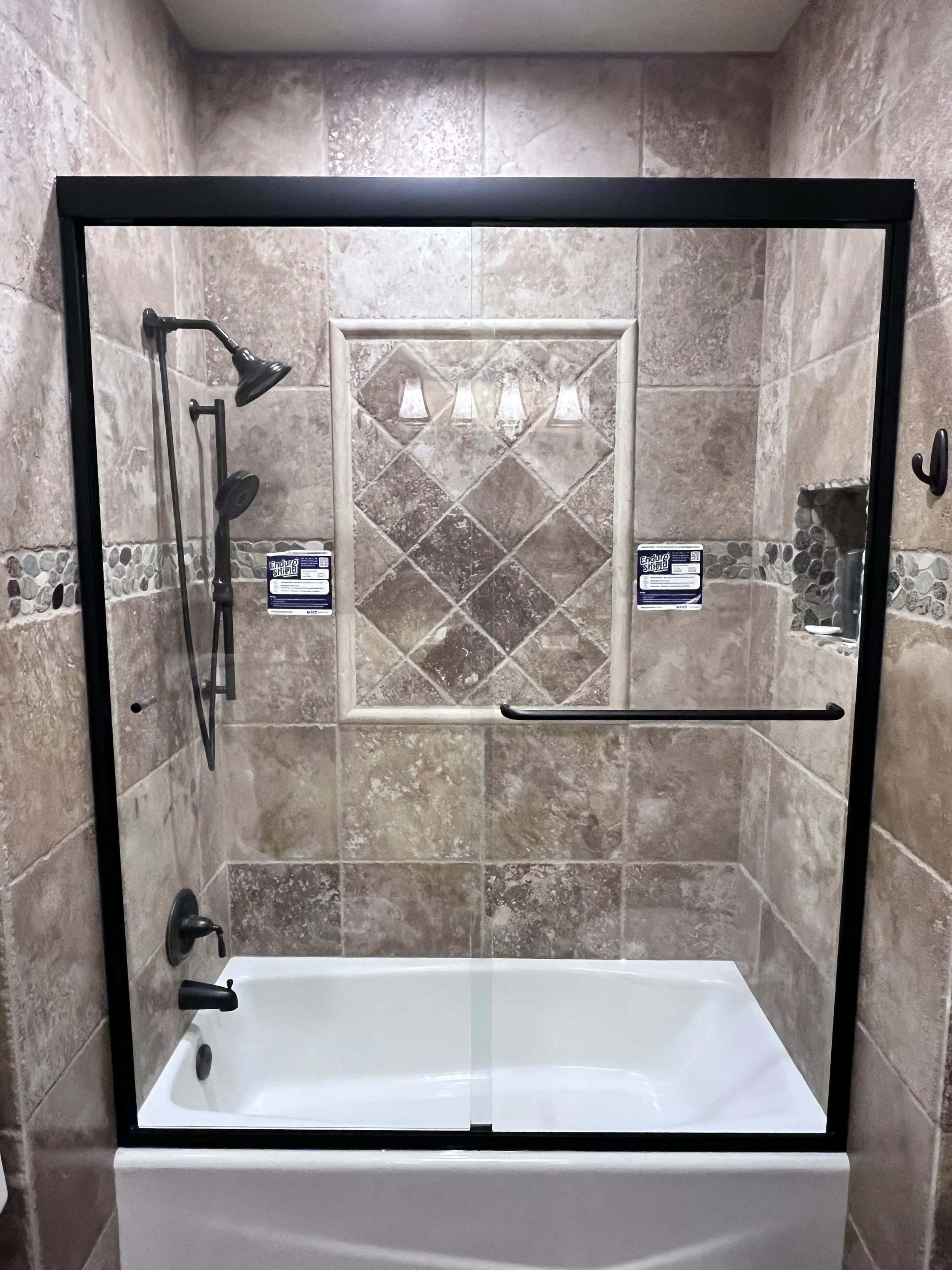 Shower with brown tiled walls, a white bathtub, a rain shower head, a handheld shower, a small built-in shelf, and a glass shower door.