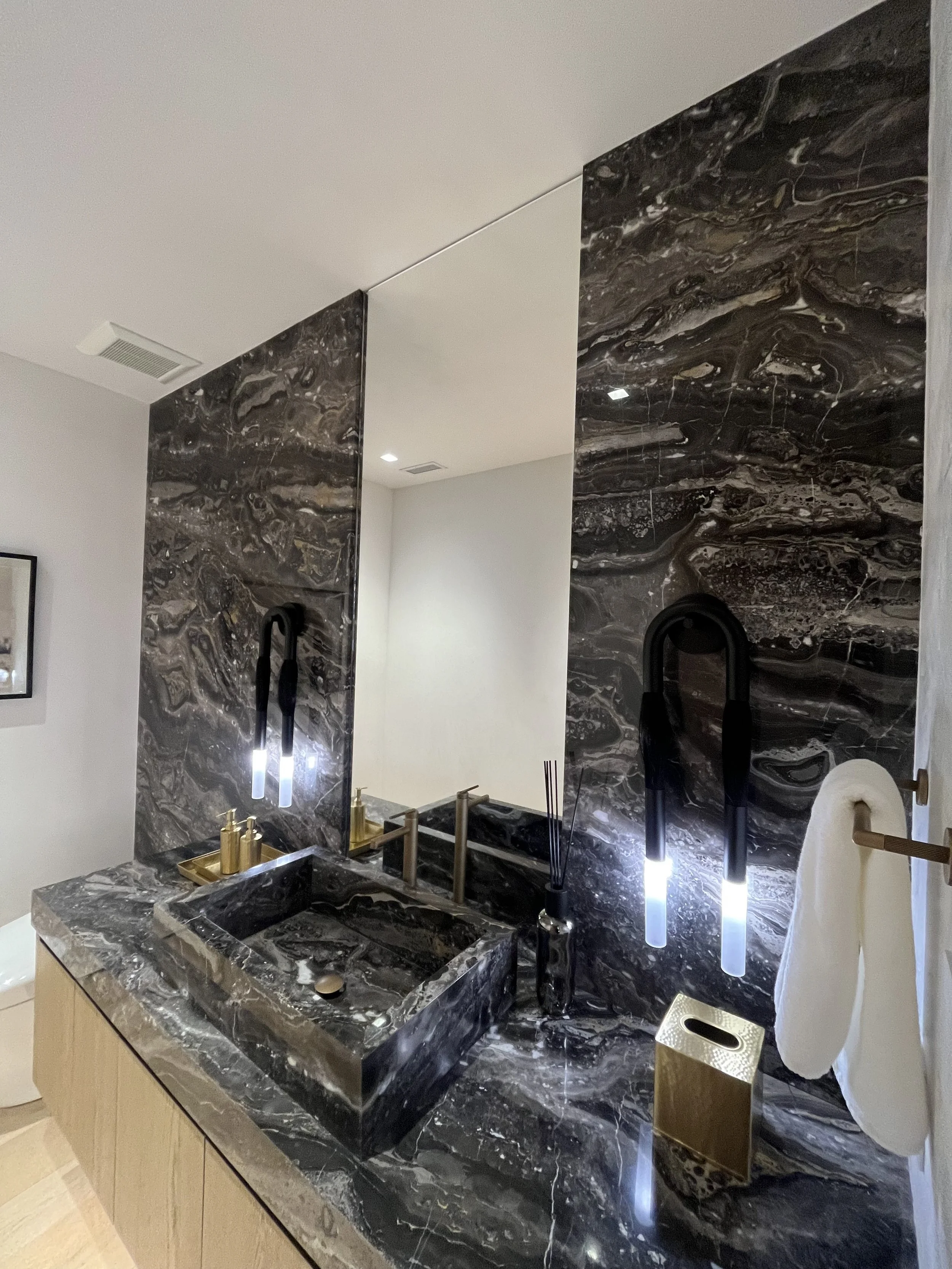 Black marble bathroom vanity with gold fixtures, black faucets, mirror, and modern black lighting, white walls, and beige floor.