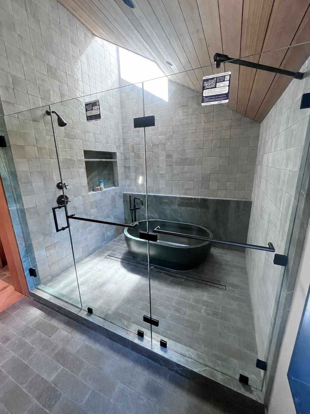 A modern walk-in shower with a glass enclosure and a dark bathtub inside. The shower has black fixtures, a built-in shelf, and a skylight above, with wooden paneling on the ceiling.