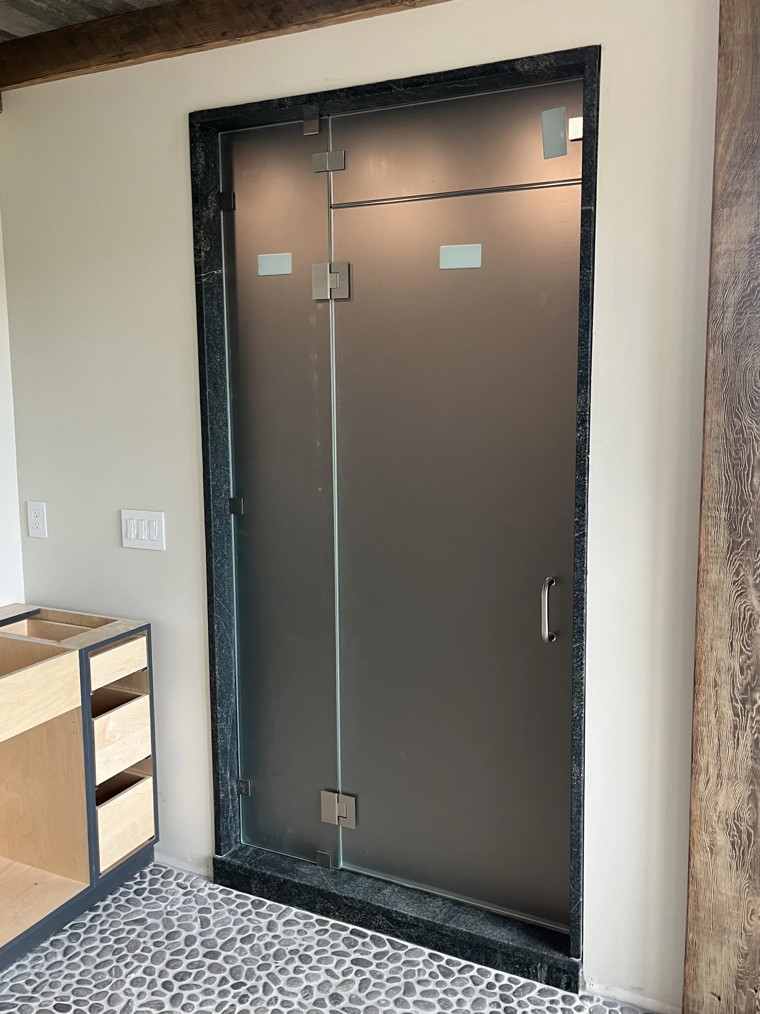 A modern, metal door with glass edges, horizontal and vertical hinges, set in a black and gray marble frame, in a room with wood accents and pebble-textured flooring.