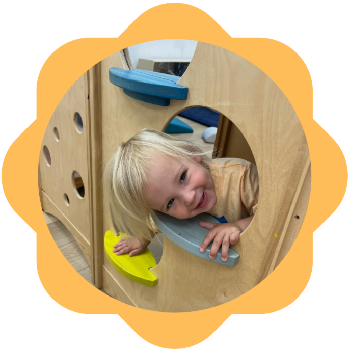 A young girl with blonde hair smiling and leaning through a circular wooden play structure with colorful elements inside.