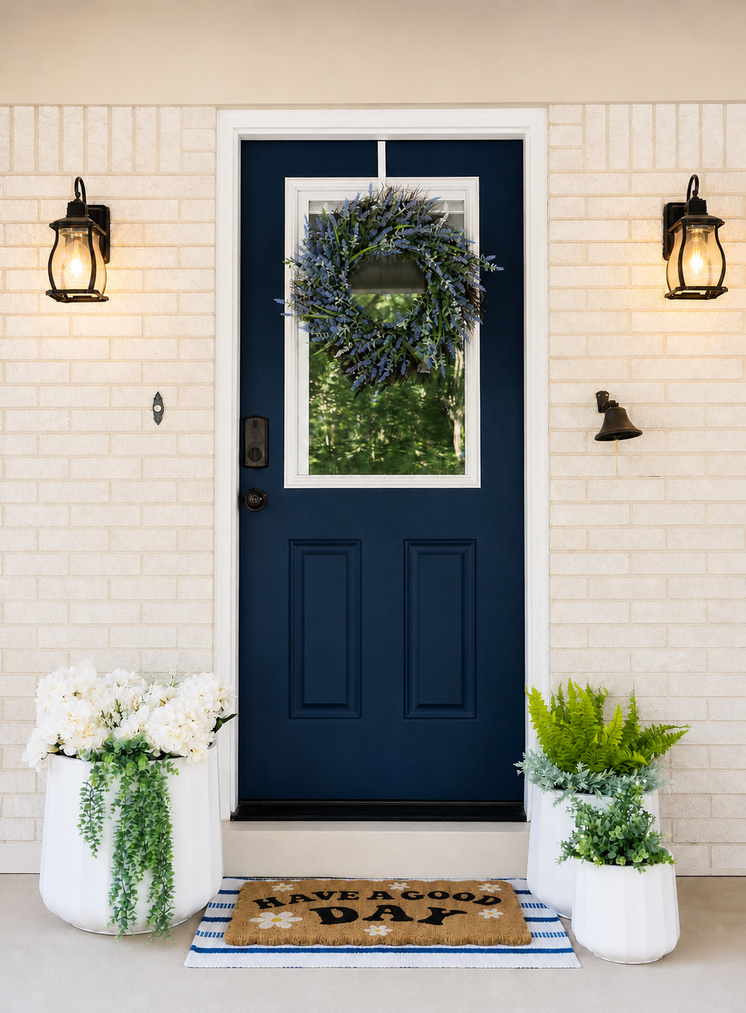 Front door decorated with a wreath, flanked by potted plants with white flowers and green foliage, and lit by two wall-mounted lanterns. 'Have a good day' doormat in front.
