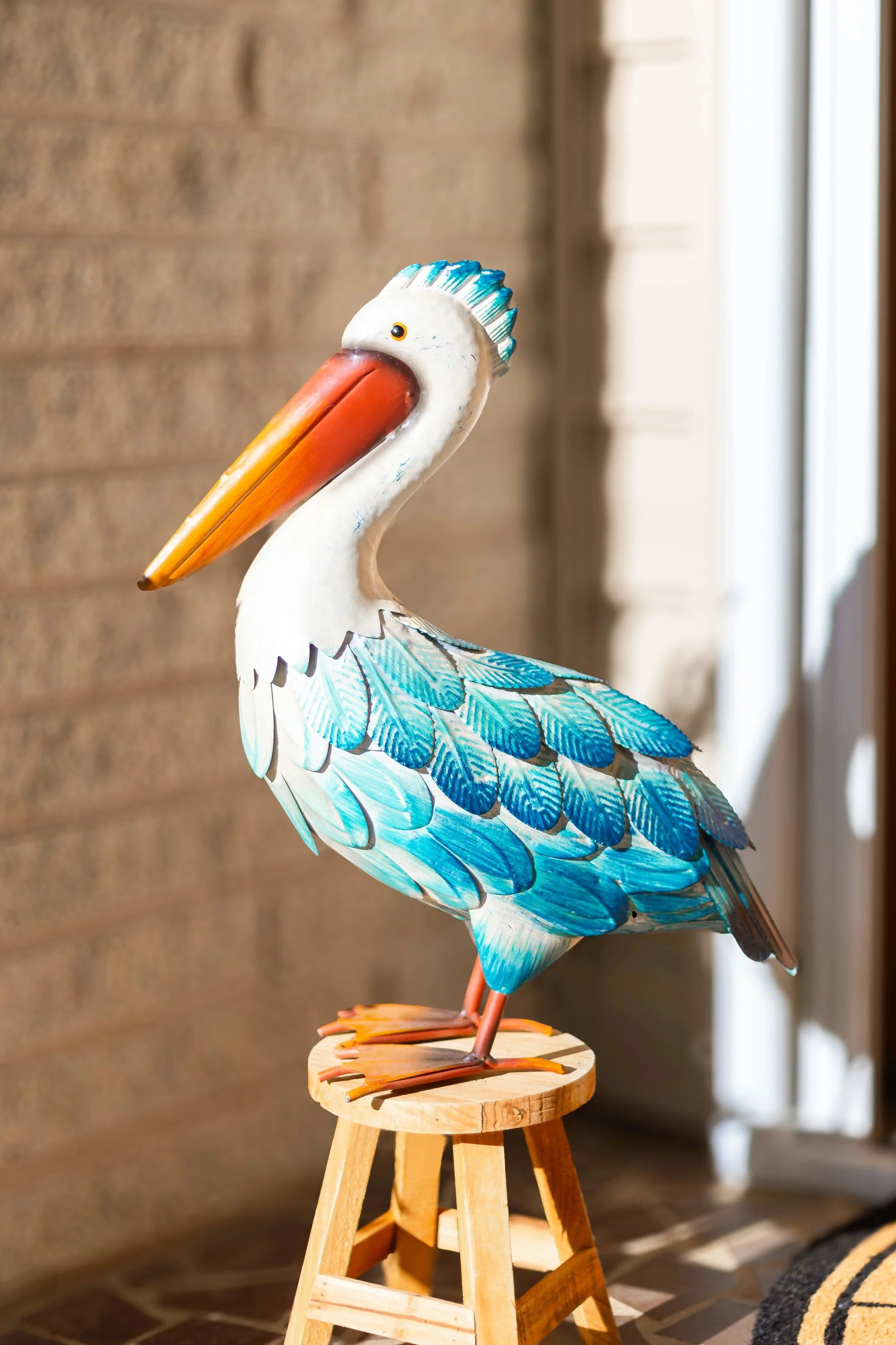 A colorful pelican sculpture with a long orange beak and blue feathers, standing on a small wooden stool.