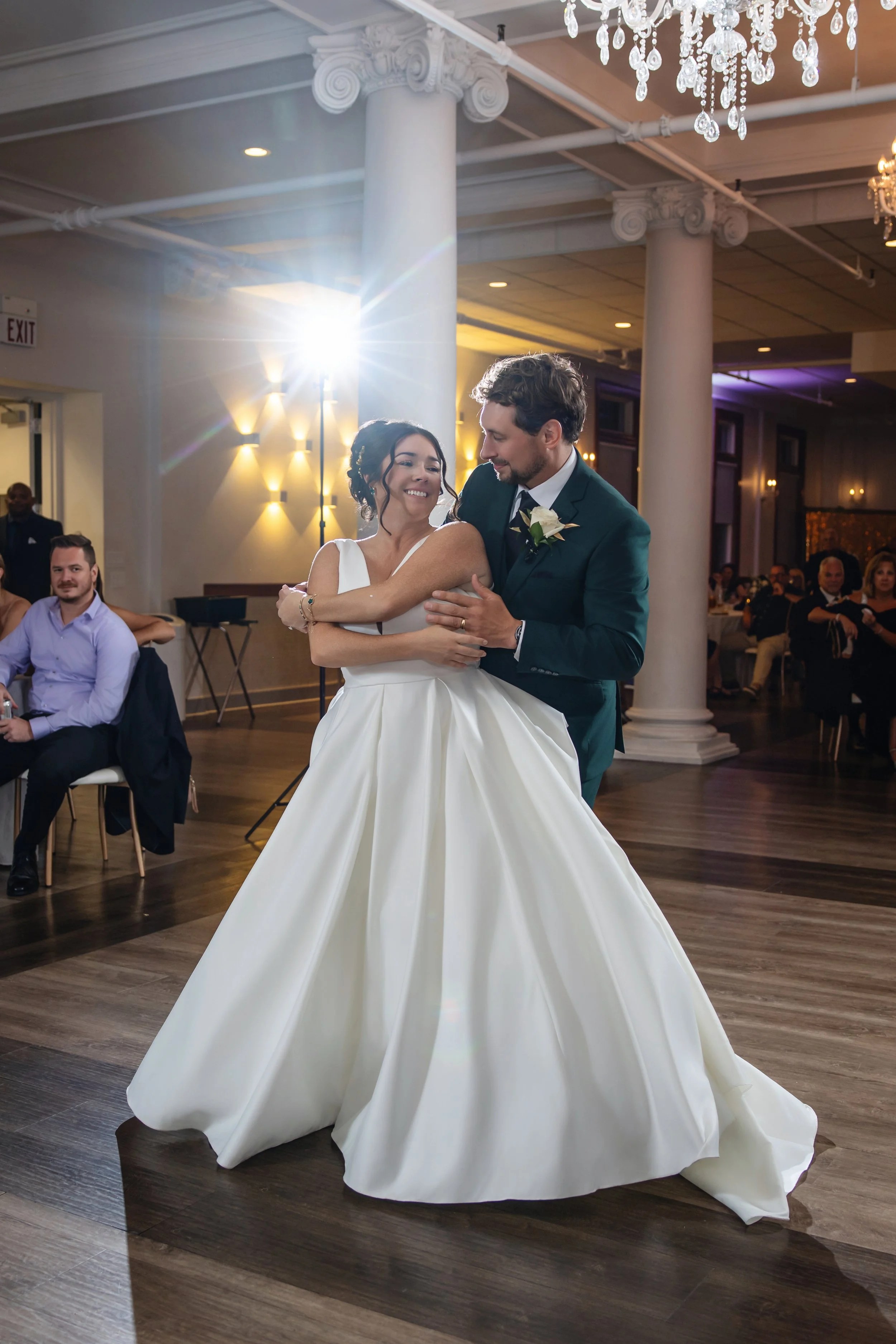 A bride and groom share a dance at their wedding reception in an elegant hall with colonial columns and chandeliers. The bride wears a white gown, and the groom sports a dark suit with a white shirt and boutonniere.