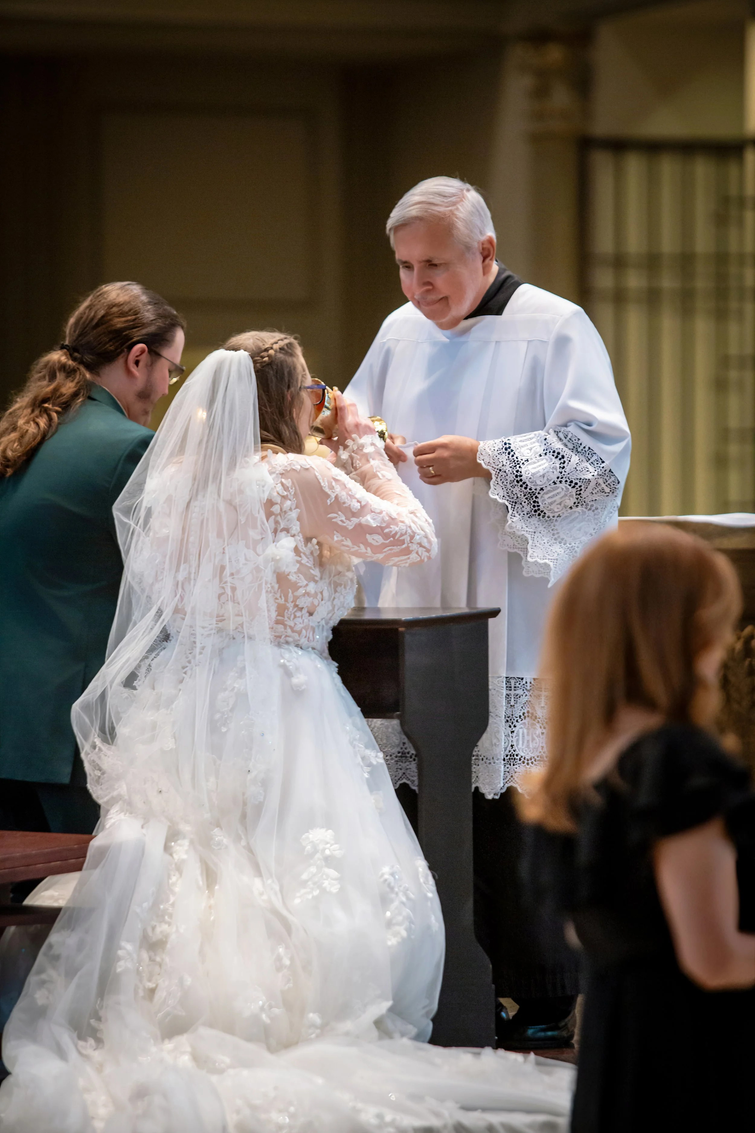 A wedding ceremony with a bride in a white lace wedding gown and veil, standing in front of a priest in an alb with lace trim, officiating the ceremony. The bride is holding the officiant's hand, with other attendees visible in the background.
