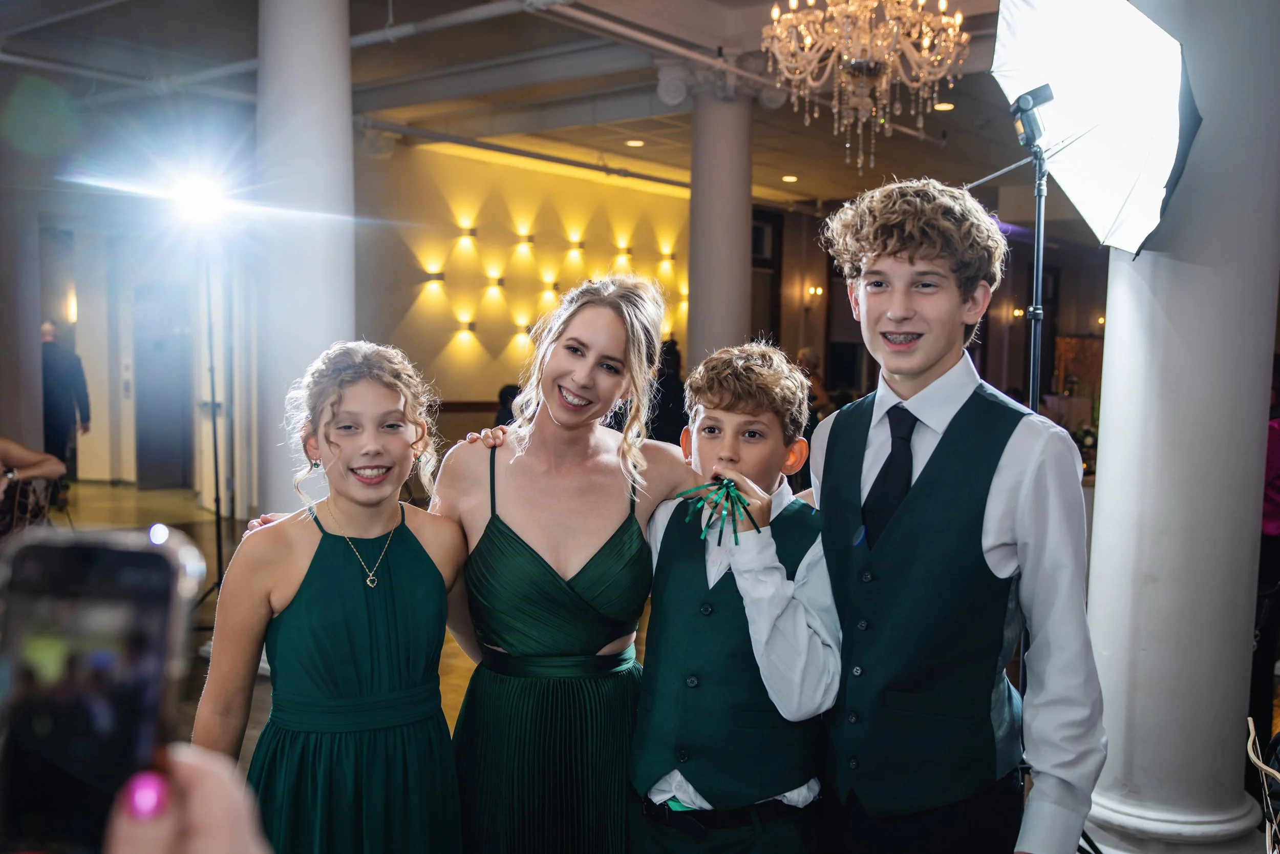 A group of four young people, including three girls and one boy, dressed in elegant green and white attire, posing together at a formal event in a decorated ballroom with warm lighting and a chandelier.