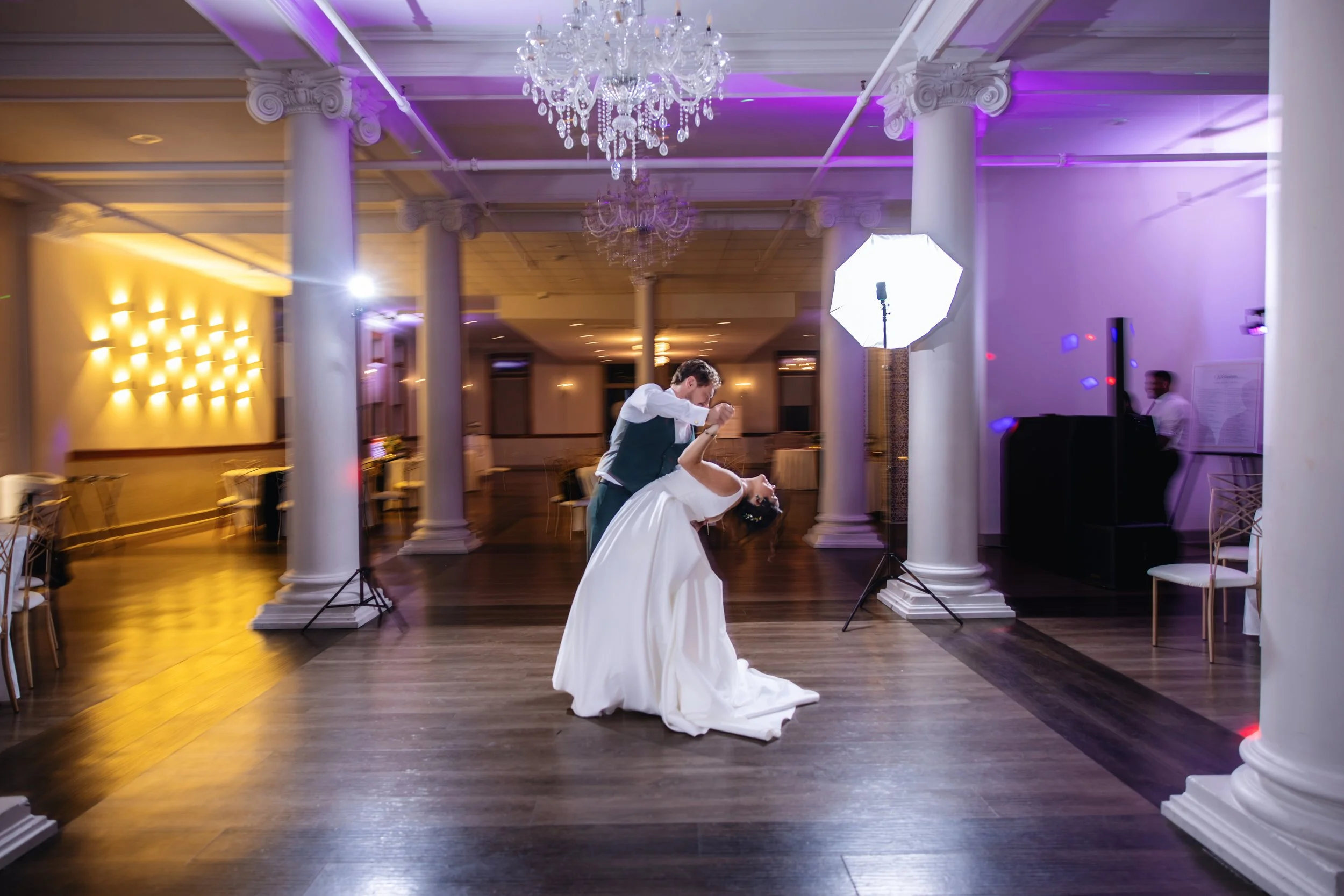 A bride and groom dancing at their wedding reception in a decorated banquet hall with chandeliers and purple accent lights.