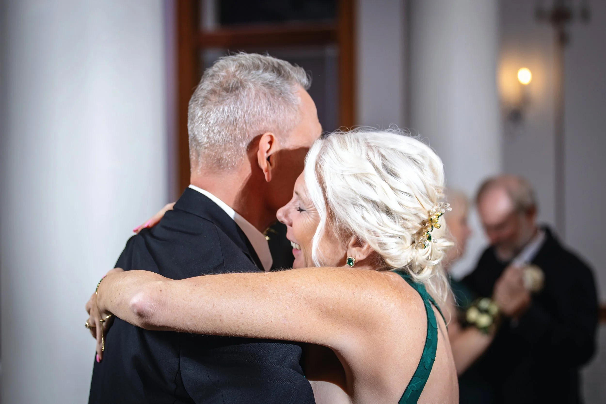 An elderly woman with white hair and green earrings dancing with a man in a black suit at a social event.