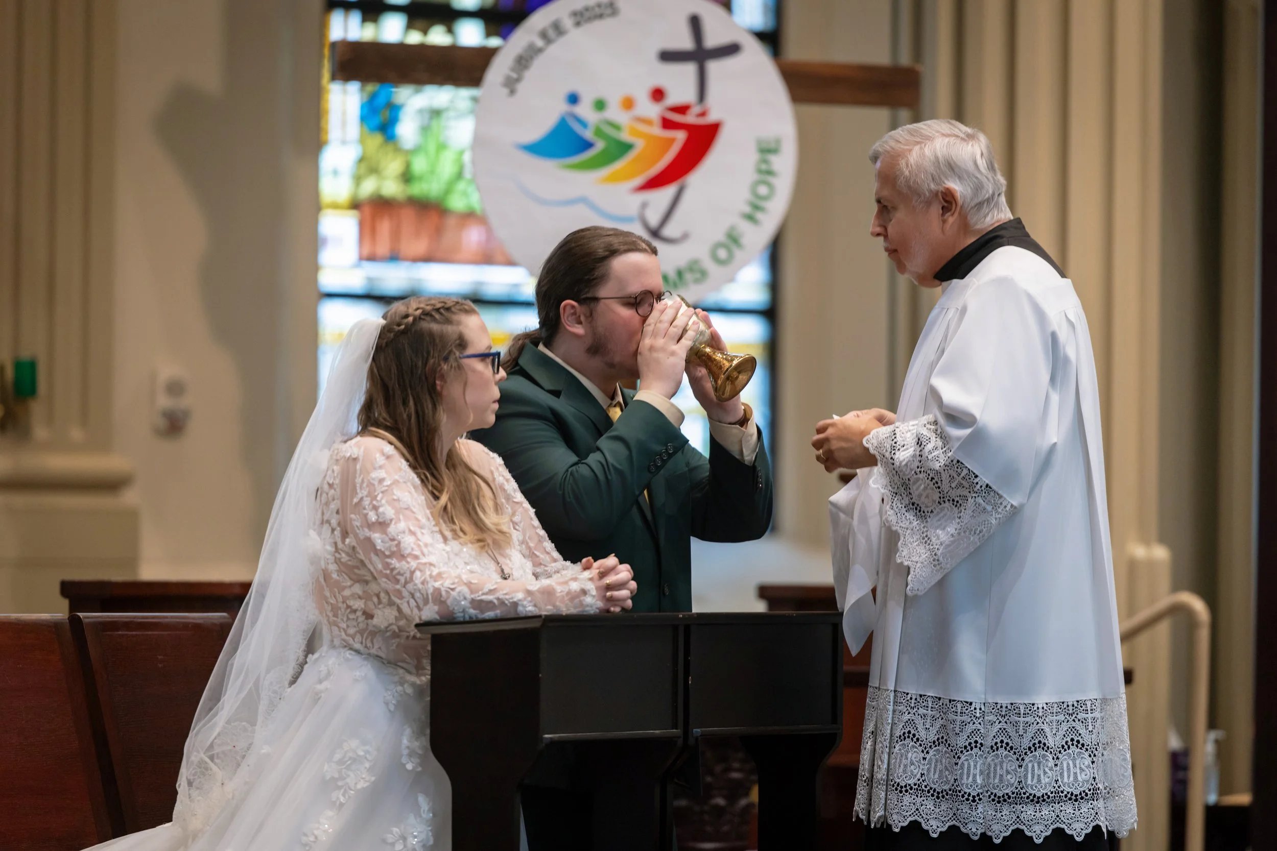 A wedding ceremony inside a church with a priest, a bride, and a groom holding a chalice. The bride is wearing a white lace wedding dress and veil, and the groom is in a dark suit. A colorful banner with a cross and dove is visible in the background.