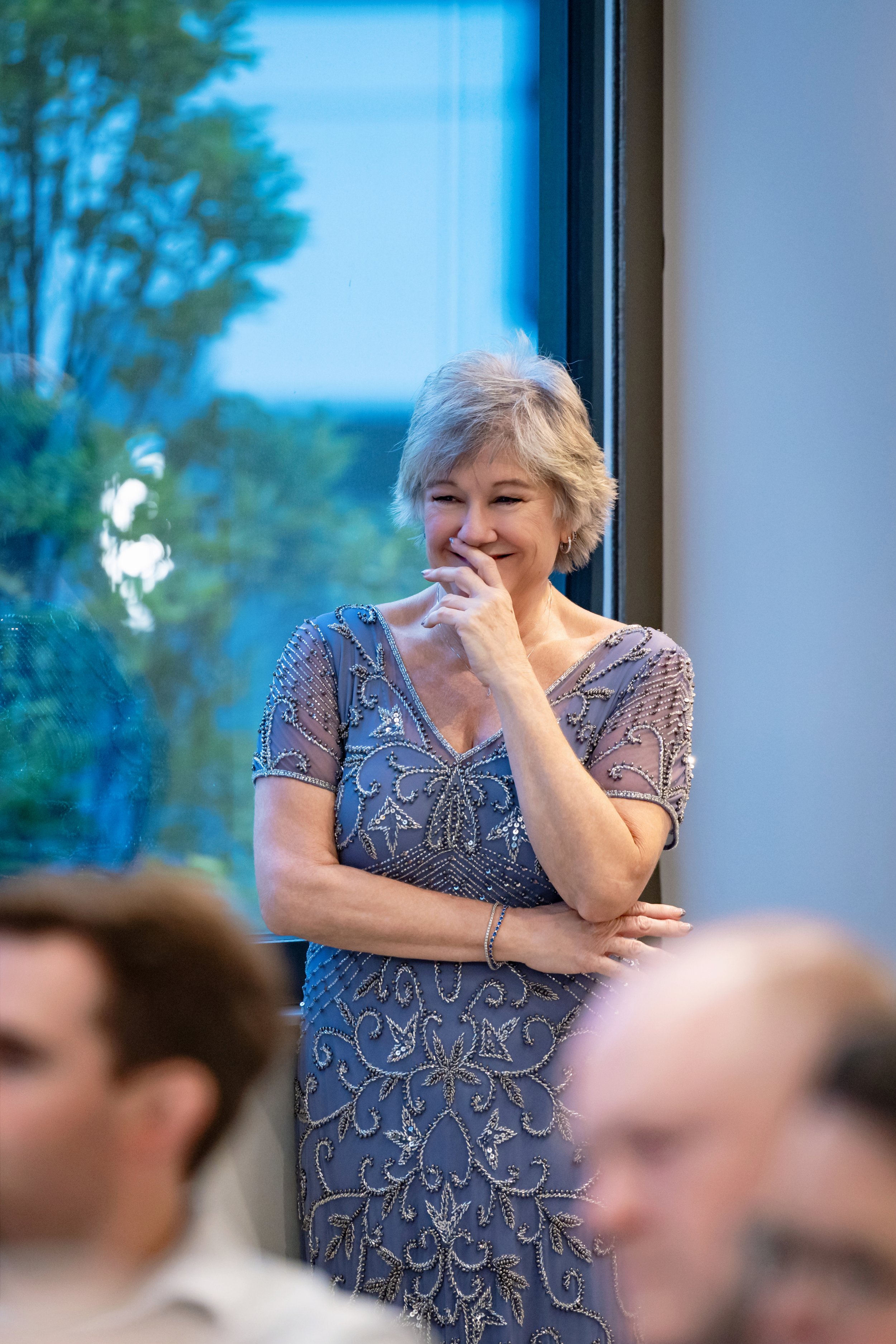 A woman in a glittery blue gown with intricate silver embroidery, smiling and touching her face, standing near a window with trees outside, at an indoor event.