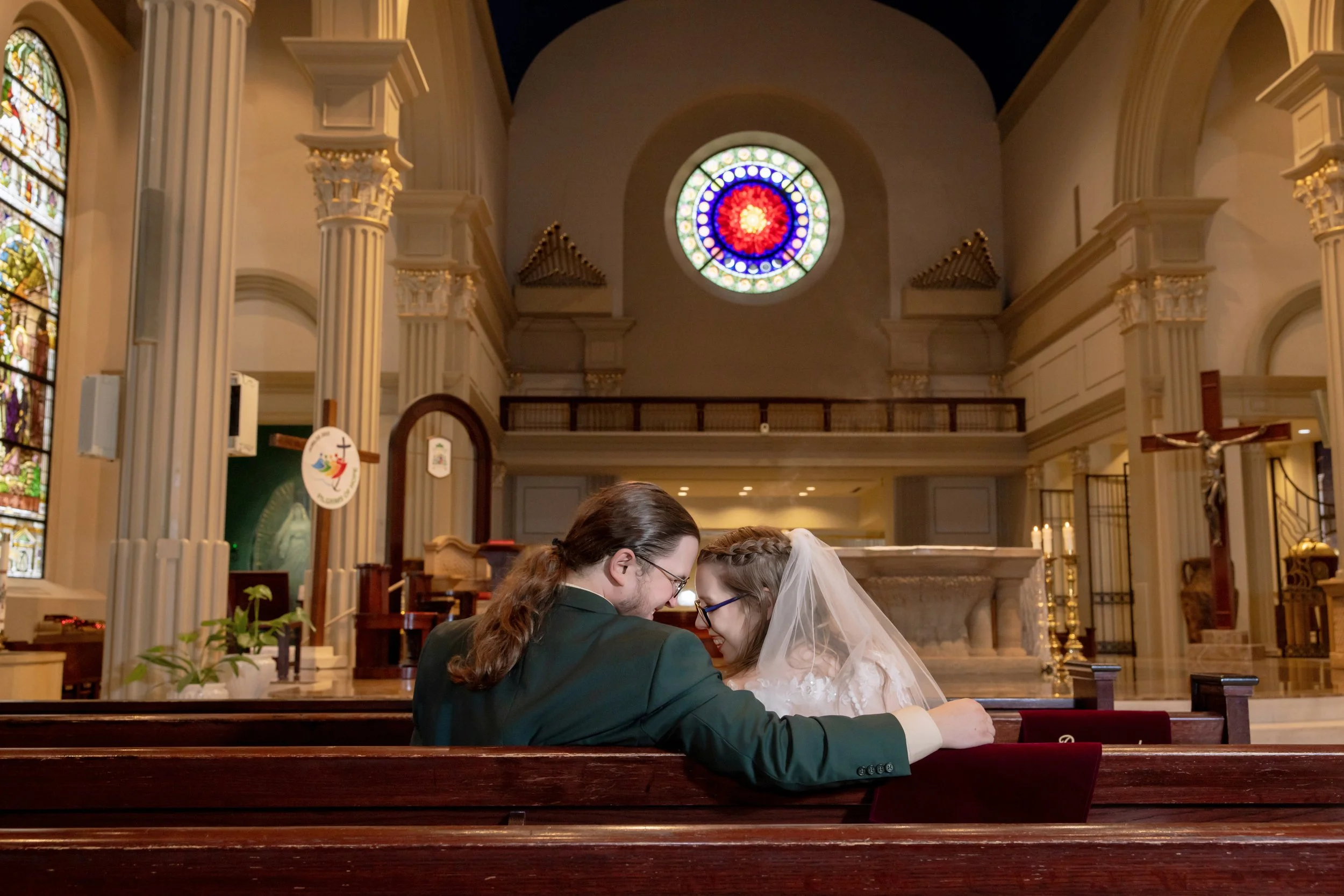 A couple getting married in a church, sitting closely with foreheads touching, wearing wedding attire, surrounded by religious stained glass windows and ornate church interior.