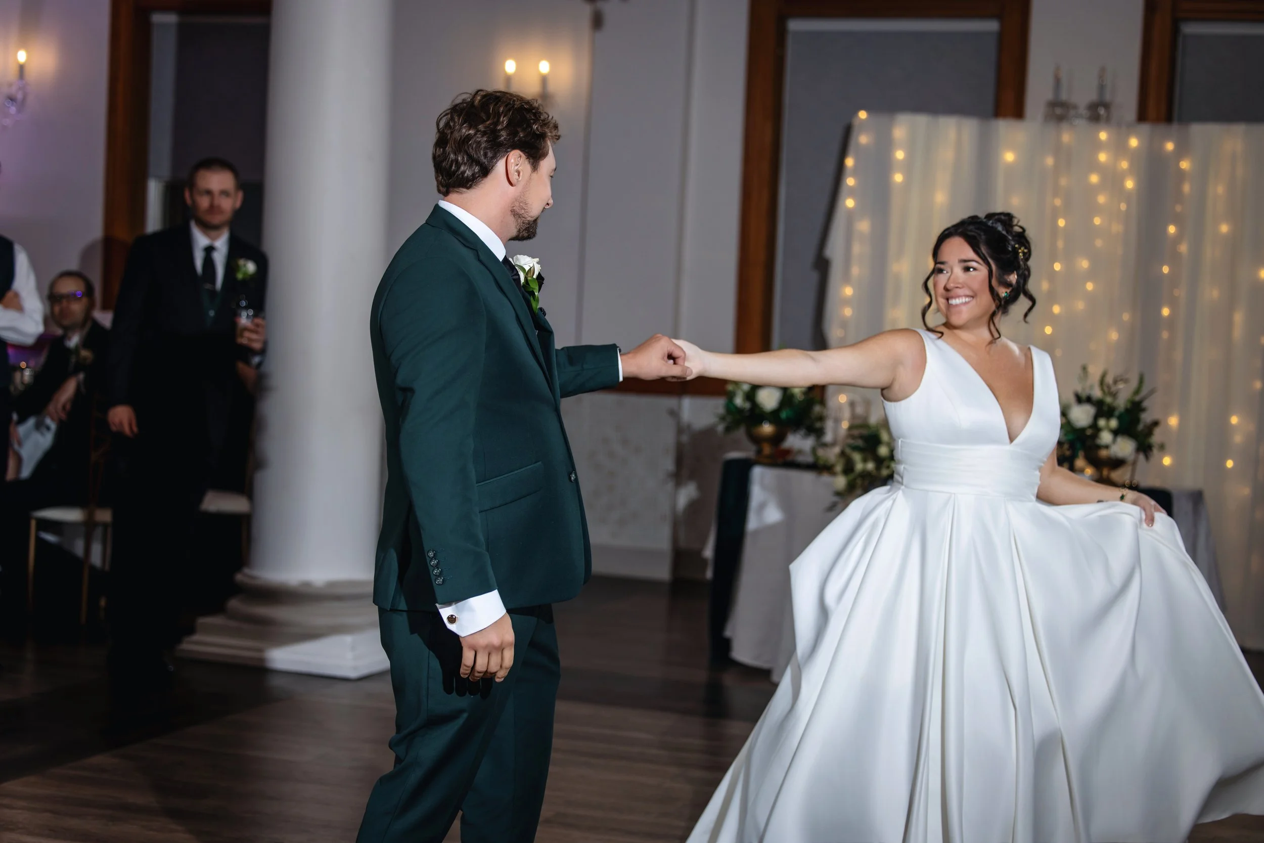 A bride and groom dance at their wedding reception, smiling at each other, with guests observing in the background.