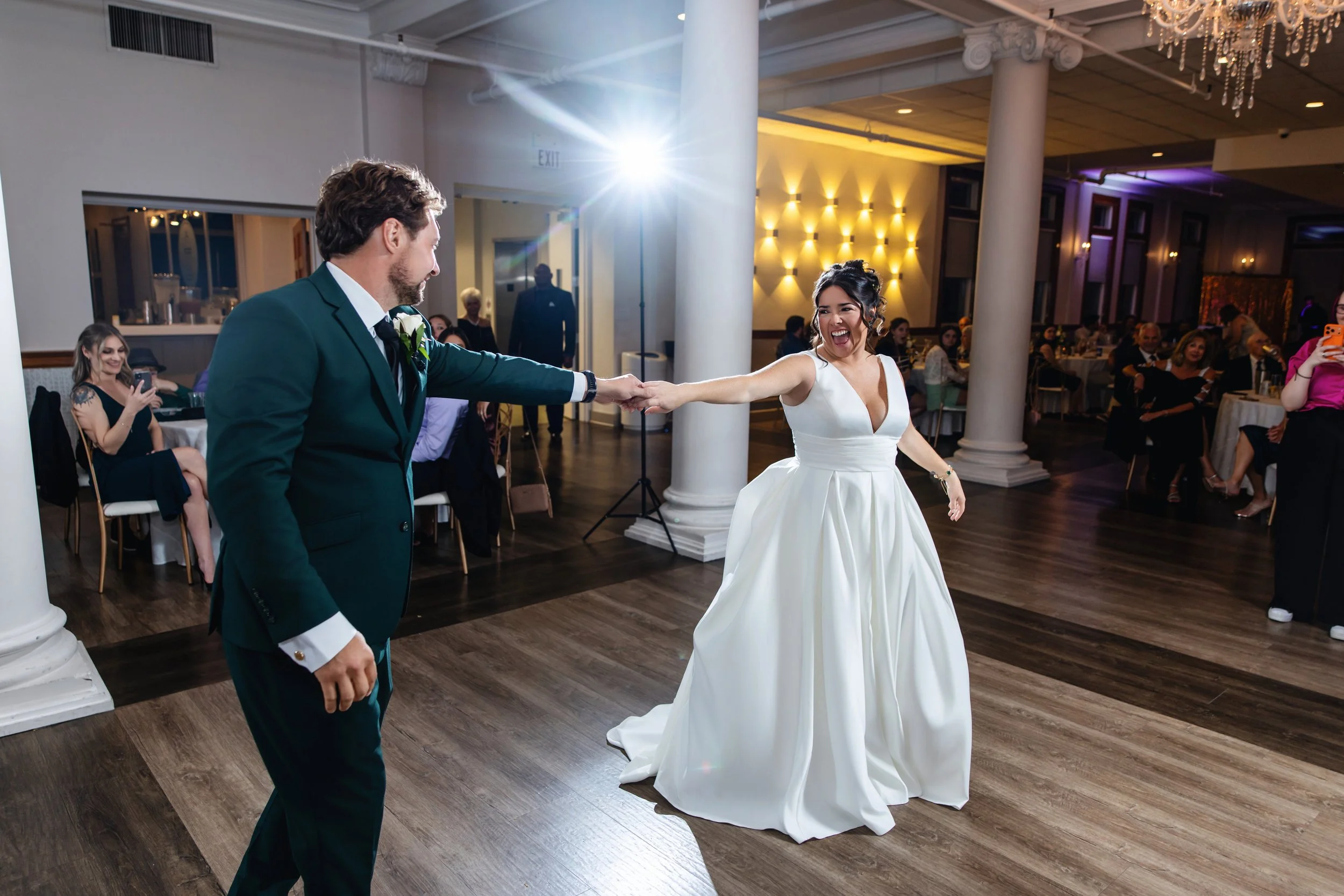 A bride and groom dance at their wedding reception, surrounded by seated guests in a decorated venue with warm lighting.