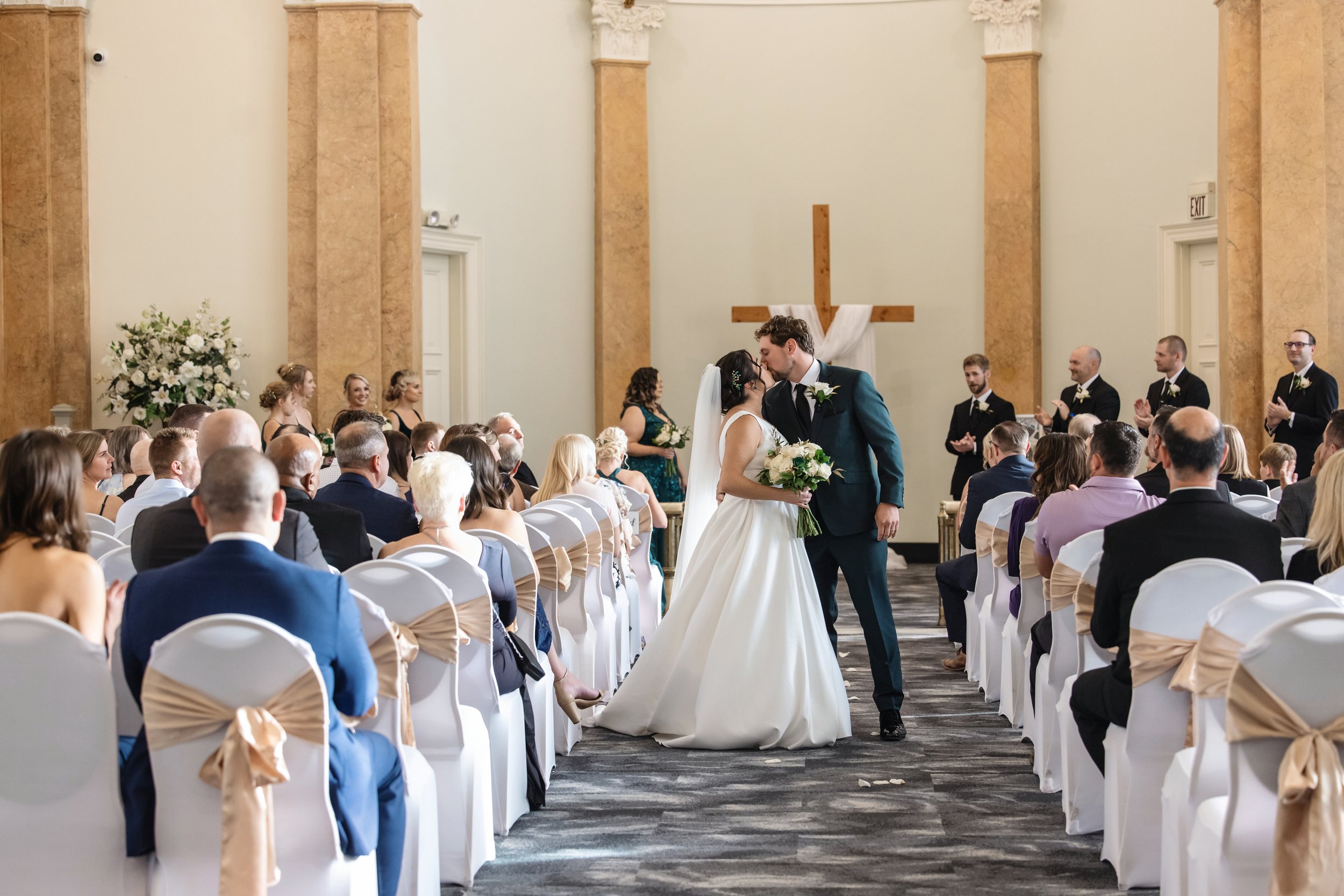 A bride and groom sharing a kiss during their wedding ceremony inside a church with a large wooden cross on the wall behind them.