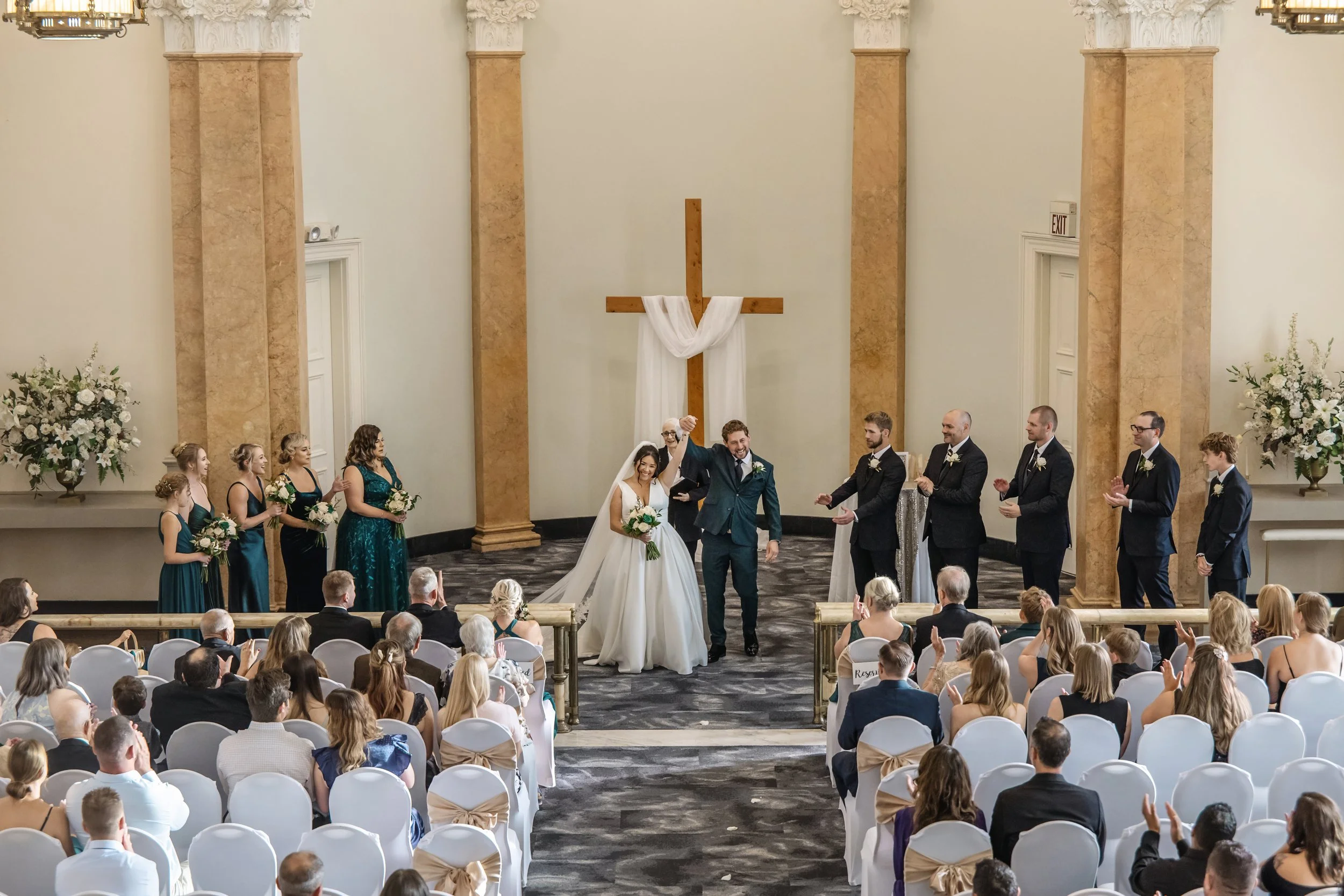A wedding ceremony taking place in a church with a large wooden cross decorated with white fabric at the altar, where a bride and groom are standing together, surrounded by bridesmaids and groomsmen, with guests seated and clapping.