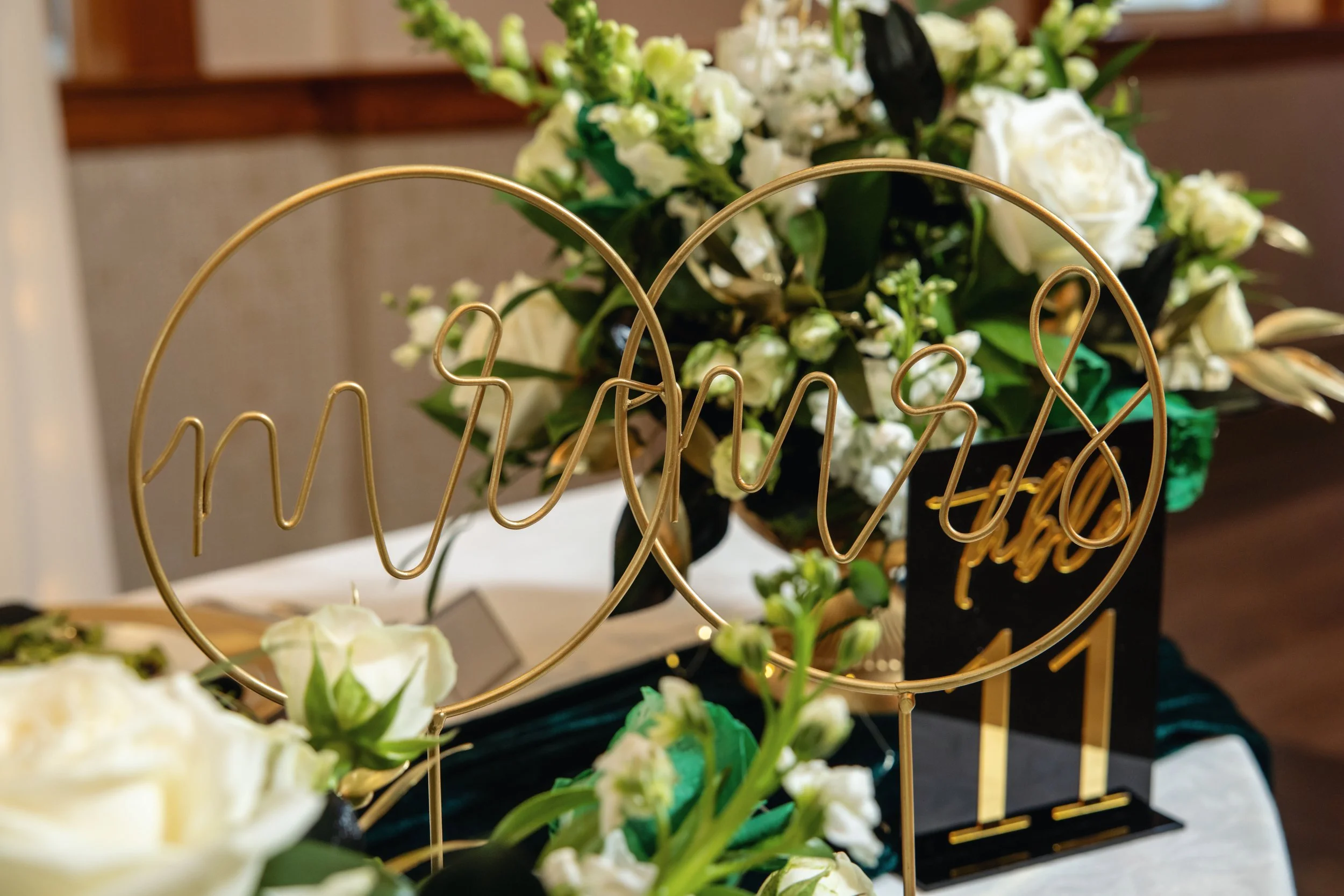 Close-up of a gold wire word sign with the word "wish" in cursive, placed among white flowers, on a white surface with a blurred background.