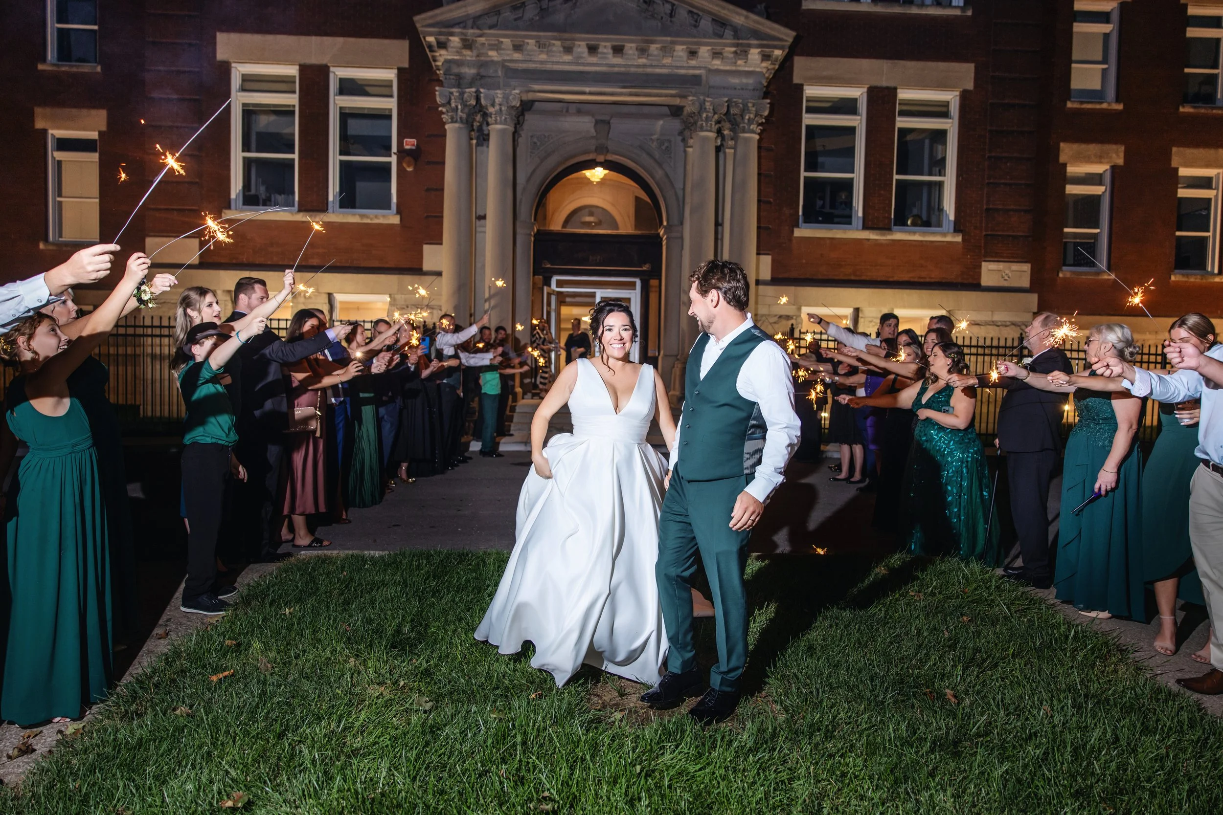 A bride and groom smile at each other during their wedding celebration outside a brick building at night, with guests holding sparklers to create a festive atmosphere.