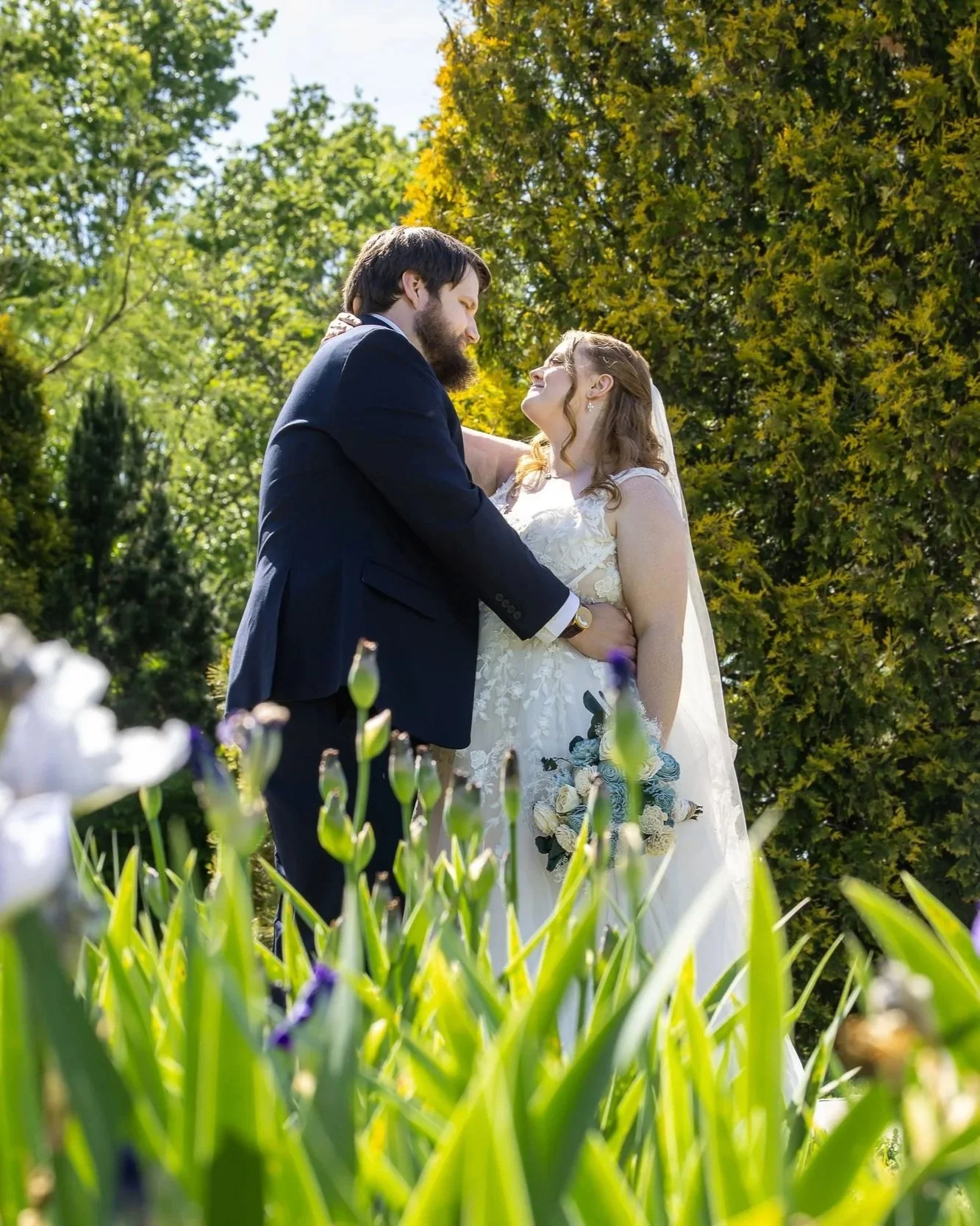 A couple gets married outdoors, standing close together amid green trees and colorful flowers on a sunny day.