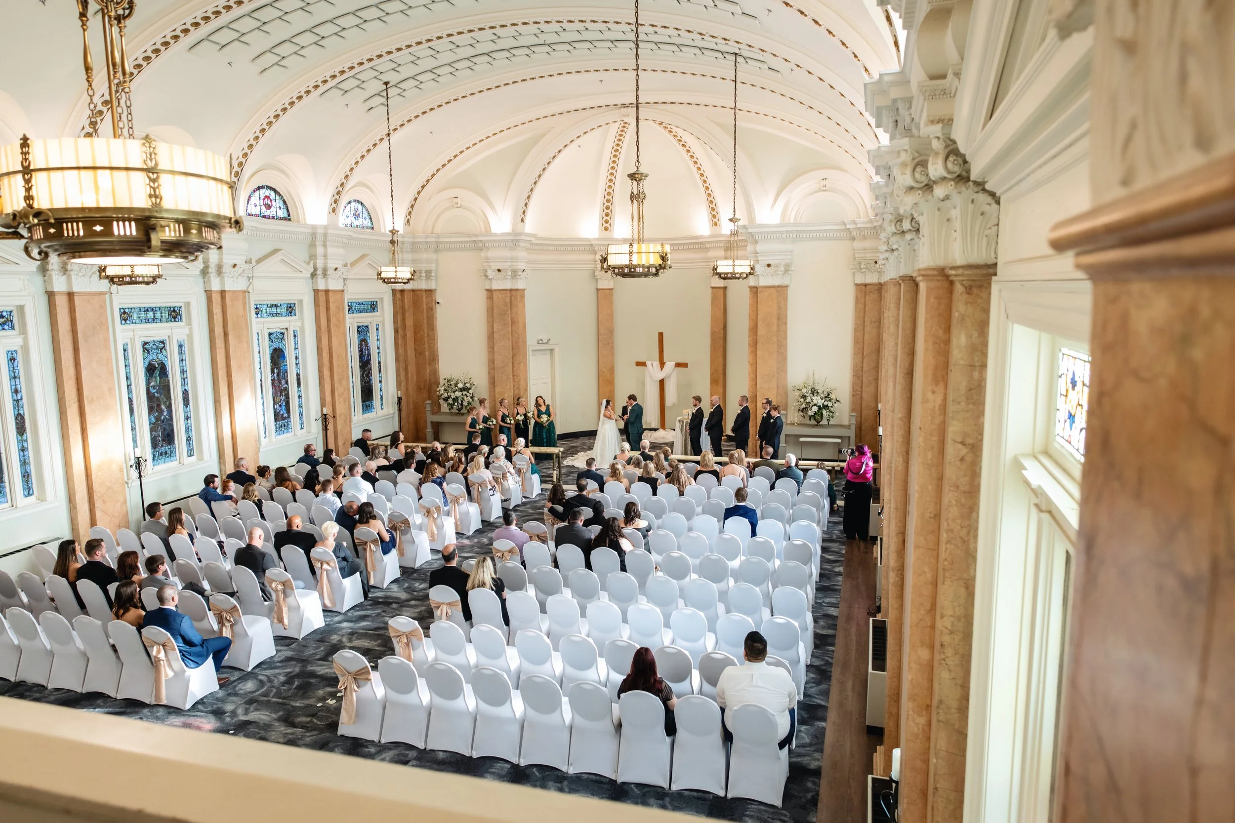 A wedding ceremony taking place inside a church with high, arched ceilings, stained glass windows, and chandeliers. The bride and groom are standing at the altar with the officiant, surrounded by bridesmaids and groomsmen. Guests seated in white chai