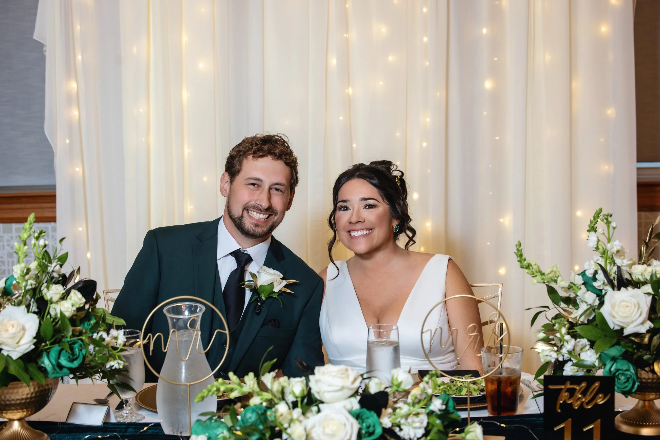 A happy bride and groom smiling at their wedding reception, sitting at a decorated table with white and green floral arrangements, surrounded by gold and glass tableware, with a soft warm background of fairy lights and cream-colored drapery.
