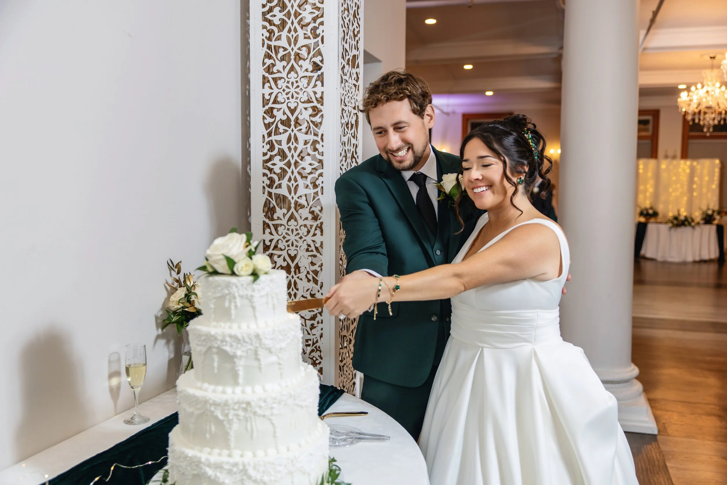 A bride and groom smiling and cutting a white wedding cake decorated with white flowers, with champagne glasses nearby, in a decorated wedding venue.