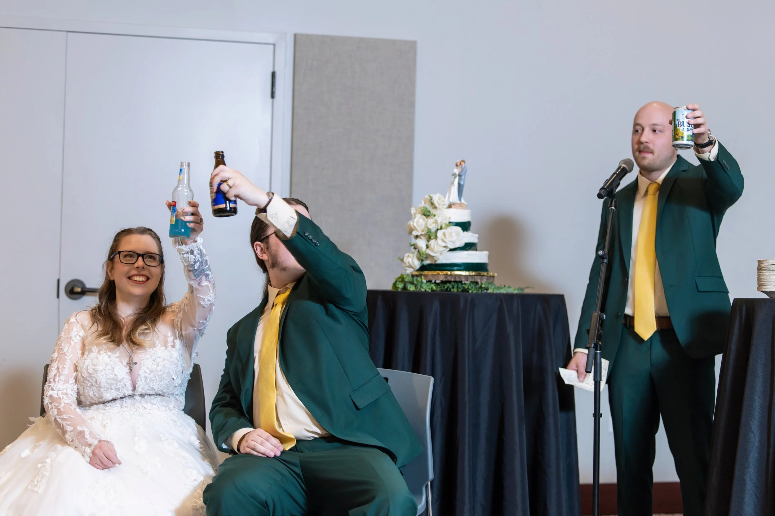Bride in a white wedding dress and glasses clinks a beverage bottle with a man in a green suit and yellow tie at a wedding reception. A three-tier wedding cake with flowers is visible in the background.