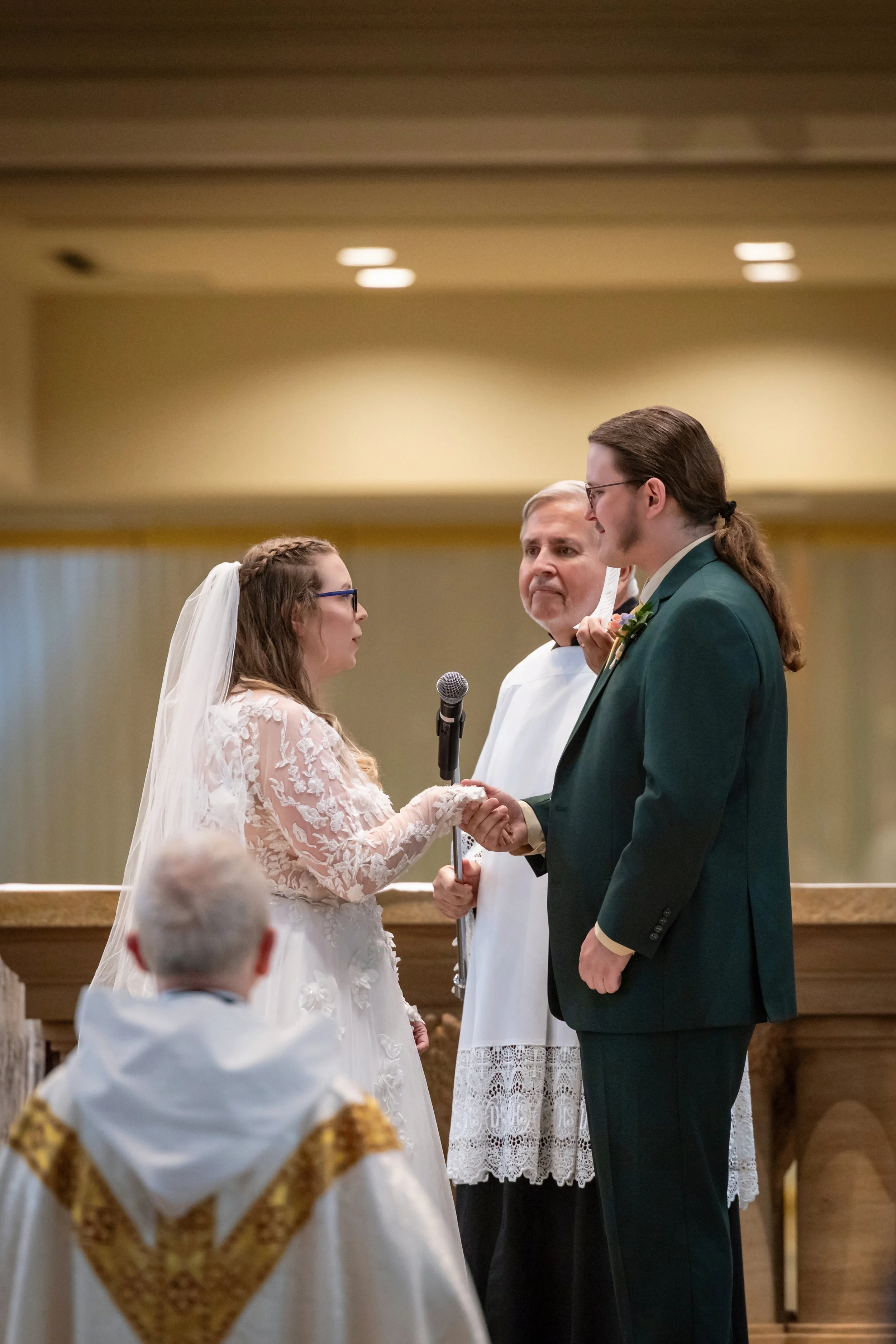 A bride and groom holding hands and facing each other during their wedding ceremony. An officiant stands between them, wearing white with lace detail. A young altar server with glasses and a white and gold robe is in the foreground. The couple appear