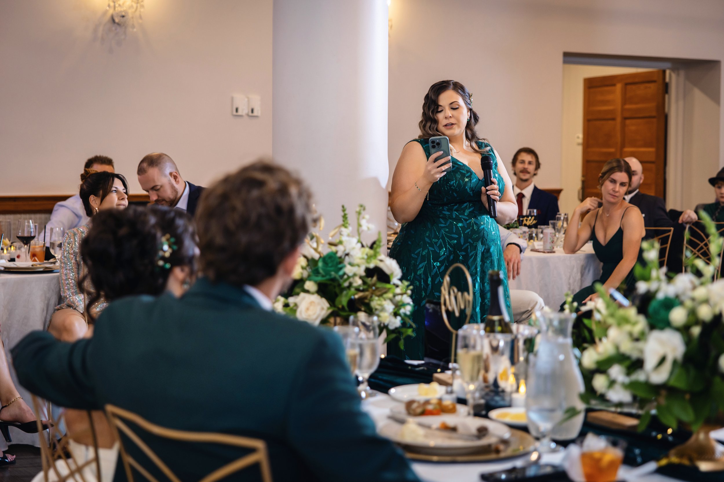 A woman in a teal dress is speaking at a wedding reception, holding a microphone in one hand and a phone in the other. Several guests are seated at tables decorated with floral centerpieces, and there are dinnerware and drinks on the tables.