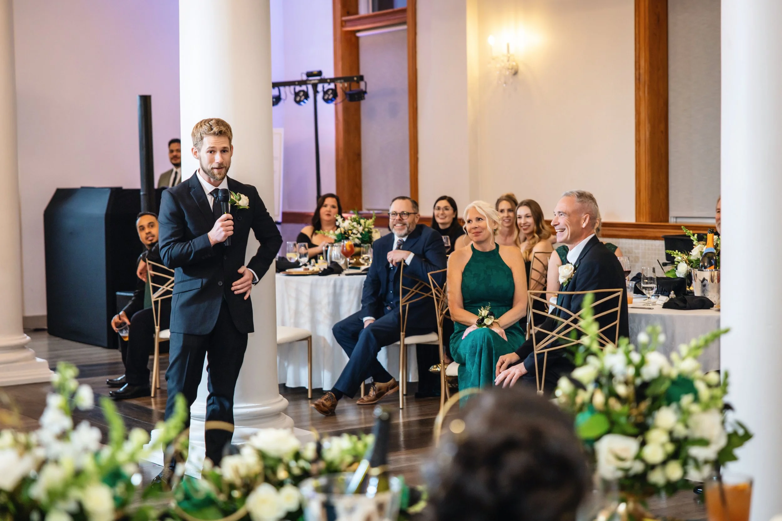 A man in a suit giving a speech at a wedding reception while guests watch and smile, decorated with floral arrangements.