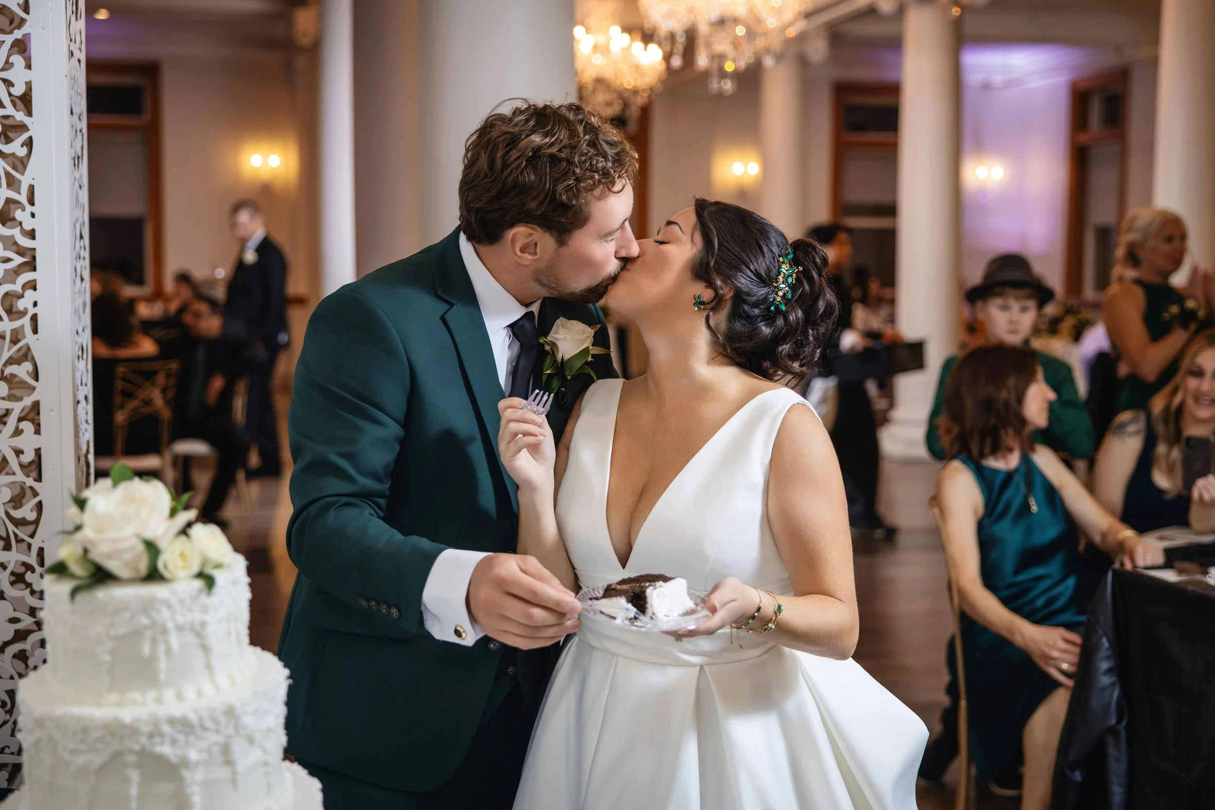 A newlywed couple sharing a kiss at their wedding reception. The groom, in a dark green suit, is holding a plate of cake, and the bride, in a white wedding dress, is holding a fork near her face. The background shows wedding guests seated at tables, 