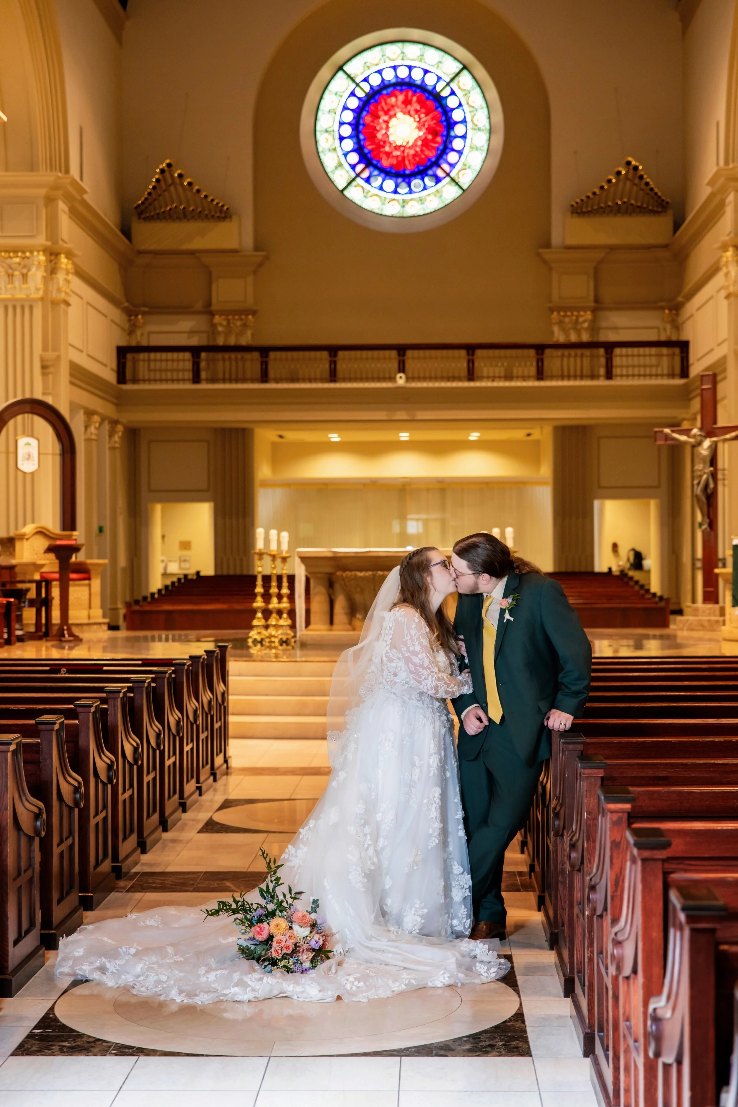 A bride and groom kissing in a church with wedding attire, floral bouquet on the floor, stained glass window above, and church pews on sides.