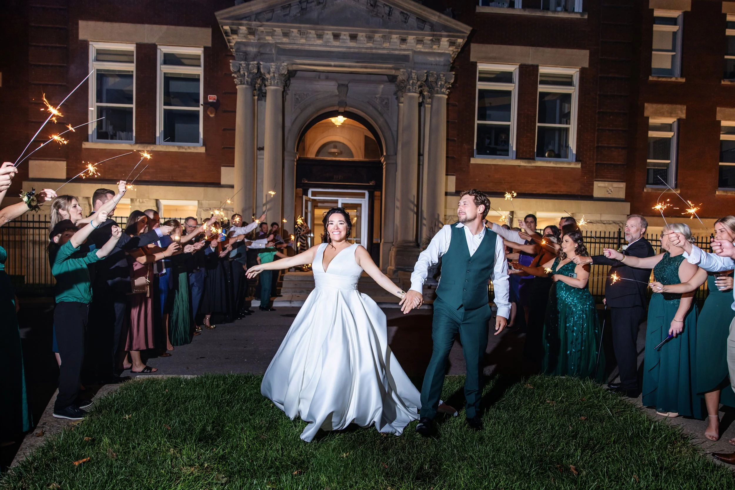 A newlywed couple holding hands and dancing in front of a line of guests holding sparklers during a nighttime wedding celebration outside a brick building with architectural columns.